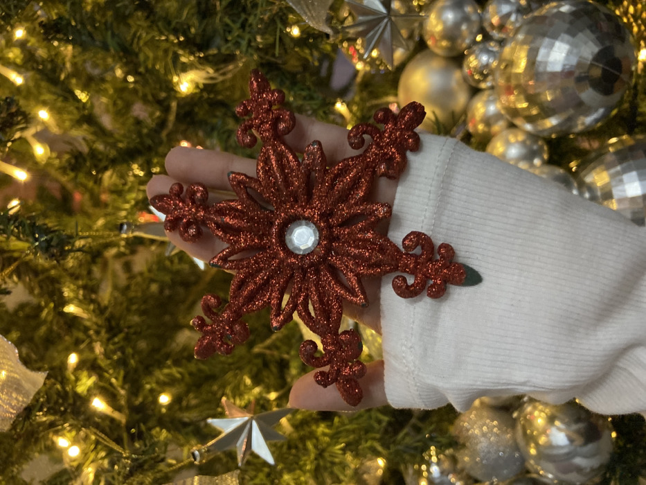 A person in a white sweater holds a sparkly, red snowflake ornament with a clear, circular gem in the center. The background showcases a Christmas tree adorned with twinkling lights, silver ornaments, and silver ribbons. The image is brightly lit and in focus, creating a festive, holiday mood.