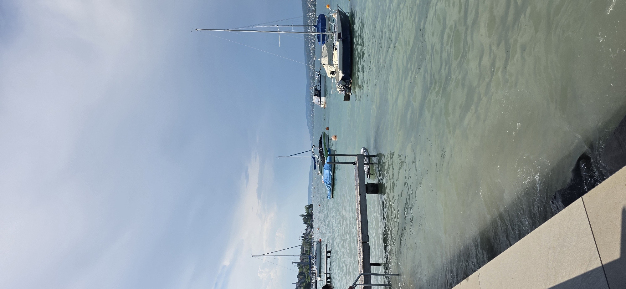The image shows a body of water with several boats anchored. A wooden pier with metal railings extends into the water from the left side of the frame. The water is calm and appears to be a lake or a large river. In the background, a city skyline is visible with buildings and trees. The sky is partly cloudy. The scene appears to be the shore of Lake Zurich in Oberrieden, Switzerland, a municipality in the canton of Zurich.