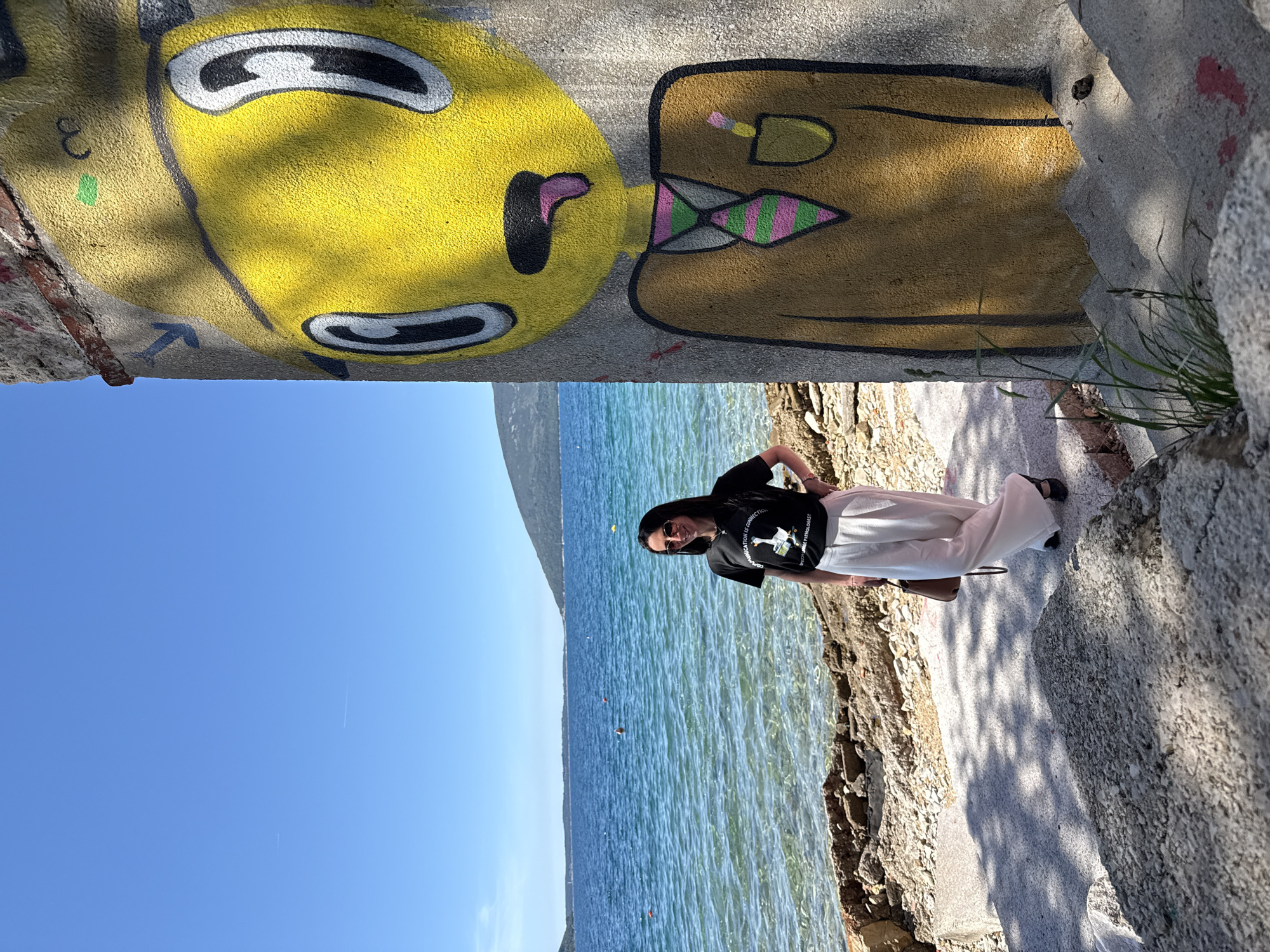 A woman in sunglasses, a black t-shirt, and white pants stands along a stone walkway beside the sea, likely in Sasovići, Montenegro, as indicated by the location. To her right is the sea, with clear blue water stretching towards a distant landmass. To her left, a concrete wall features yellow graffiti art. The sky above is clear blue.