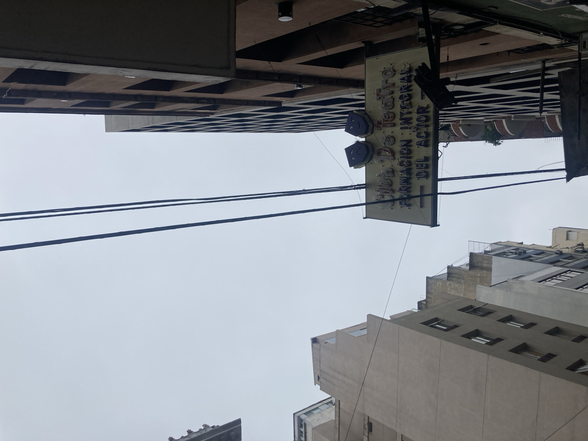 The image captures an urban street scene dominated by tall buildings under an overcast sky. The photograph appears to have been taken looking upwards, with the main subject being a rectangular sign hanging perpendicularly from a building.
The sign is a light yellow or cream color with dark blue text and graphics. It features the words "CLUB DE TEATRO" at the top, followed by "FORMACIÓN INTEGRAL" and "DEL ACTOR" below. These phrases translate from Spanish to "Theater Club," "Comprehensive Training," and "OF THE ACTOR," indicating an institution related to performing arts. Attached to the sign, beneath "CLUB DE TEATRO," are two stylized masks, likely representing the traditional theatrical symbols of comedy and tragedy. Electrical or utility wires stretch horizontally across the middle of the frame.
The buildings surrounding the sign are modern, multistory structures with various architectural details, including prominent window patterns and layered facades. Some buildings appear to be made of concrete or similar materials in shades of gray and beige. The sky is uniformly light gray, suggesting an overcast day with no direct sunlight. No people or specific activities are visible in the frame, emphasizing the architectural and urban landscape.