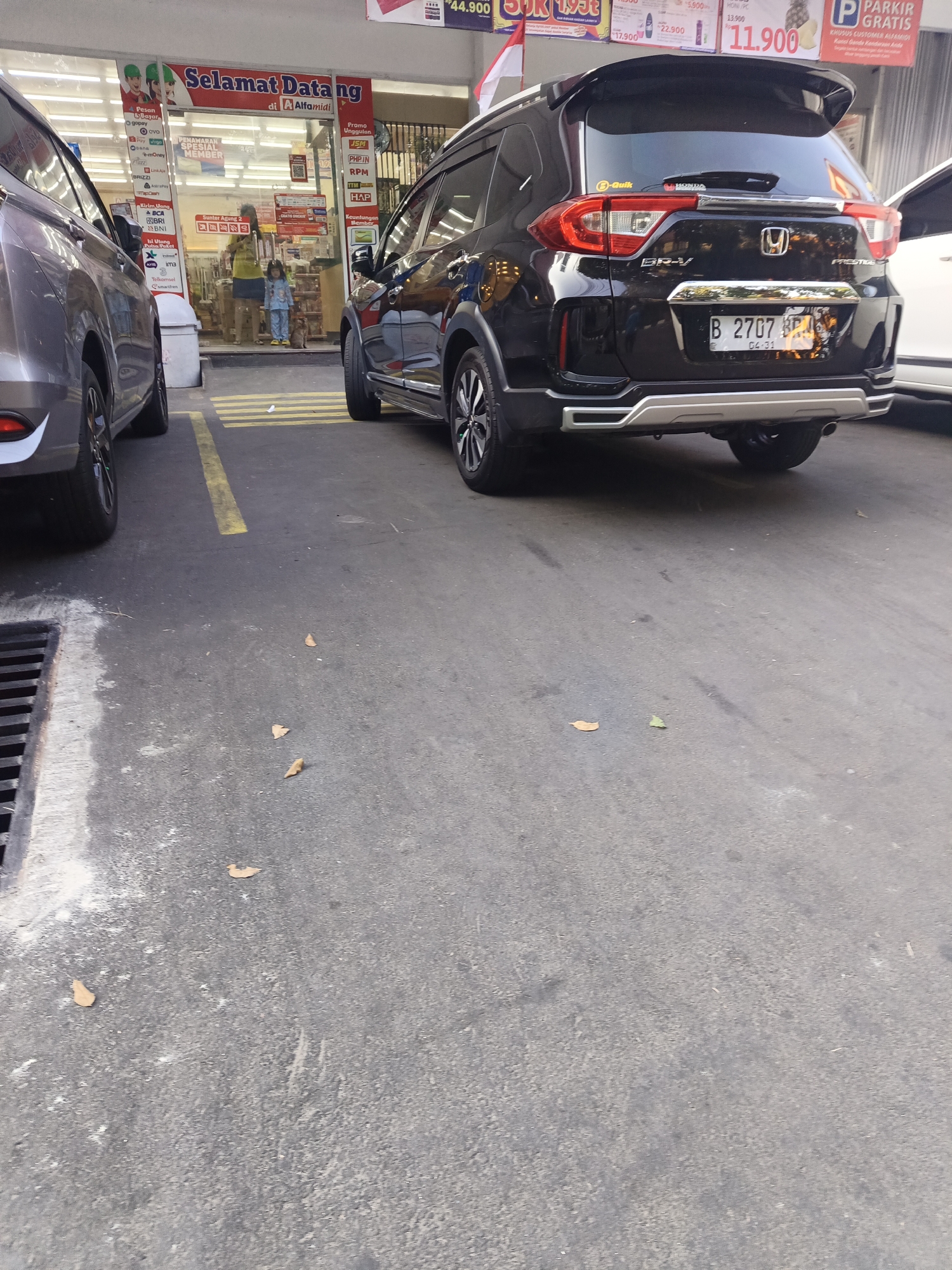 Two cars are parked in front of an Alfamidi convenience store in North Jakarta, Indonesia. The car on the right is a black Honda BR-V Prestige. It has license plate number B 2707 KPA. The car on the left is a gray SUV. The store entrance is visible behind the cars, with signs in Indonesian. A woman and a child are standing inside the store. The ground is paved asphalt. There are yellow lines painted on the ground to mark parking spaces. A drain is visible on the left side of the image.