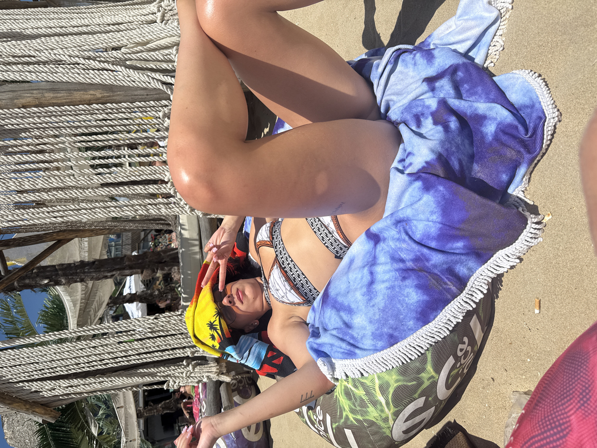 A young woman lies on a patterned beanbag chair on a sandy beach. She is wearing a bikini and sunglasses, with a colorful towel draped over her head and shoulders. Her right arm is raised, and her fingers are posed in a peace sign. The background shows part of a beach bar or restaurant structure with rope detailing and palm trees. The scene is lit by bright sunlight. The location is identified as Nesebar, Bulgaria.