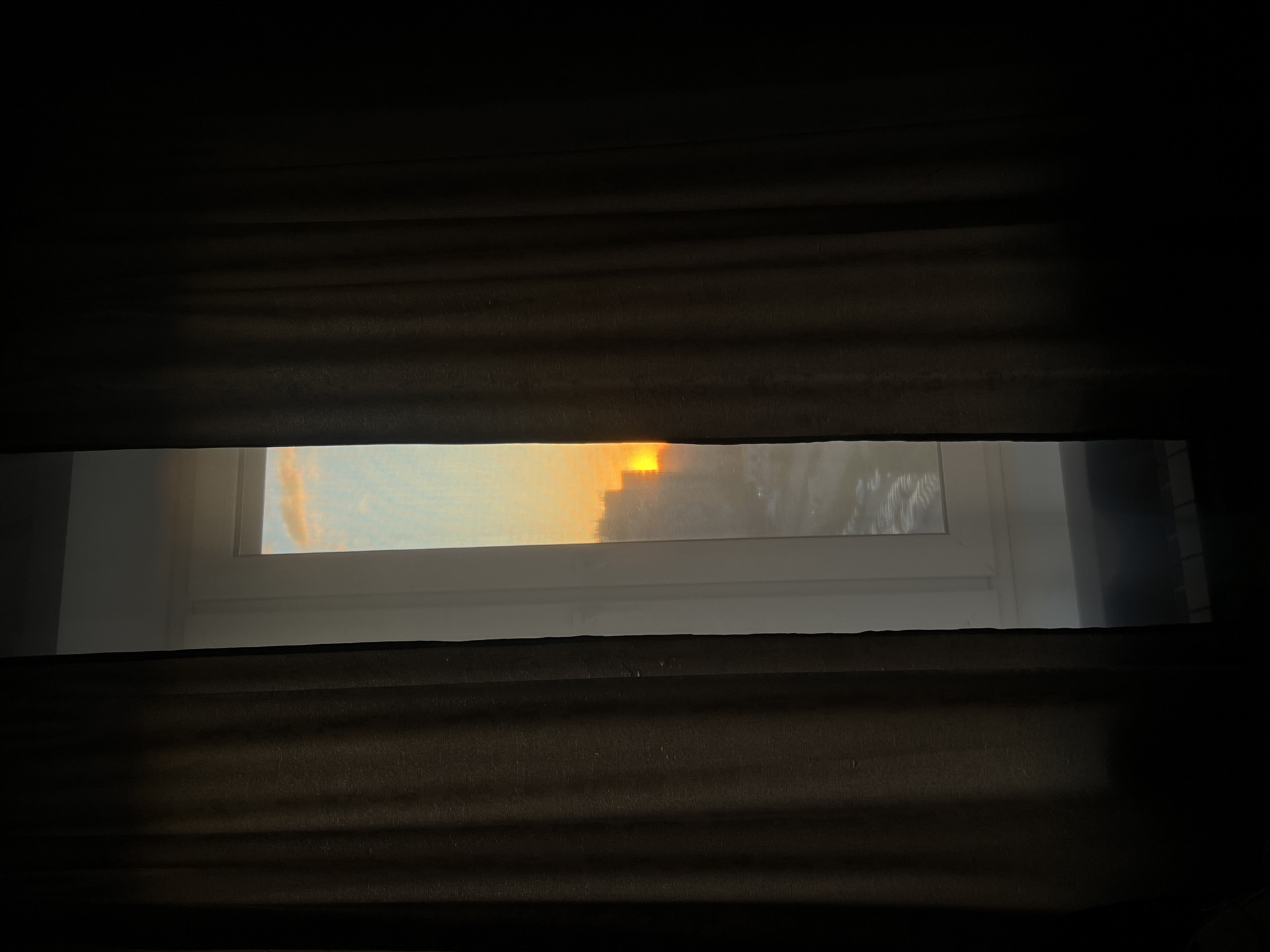 An interior shot captures a vertical segment of a window in Dachnoe, Russia, framed by two dark, heavy curtains drawn mostly closed. The curtains, dark brown with discernible vertical pleats and texture, occupy the majority of the left and right sides of the image. A white window frame is visible in the central opening. Through this aperture, the sky at what appears to be sunrise or sunset is visible, presenting a light blue hue with subtle orange and pink cloud formations in the upper section. Below the sky, silhouettes of multiple distant buildings are apparent, partially obscured by atmospheric effects. A bright, intense orange light source, presumed to be the sun, is positioned slightly to the right of the vertical center within the visible window area, casting a strong glow. Beneath the distant structures, a wide, reflective surface, possibly a body of water or a road, is present, displaying faint patterns indicative of ripples or textures. The lowest visible part of the exterior view is partially obscured by a dark interior element, potentially a windowsill. No individuals, animals, or explicit actions are observed.