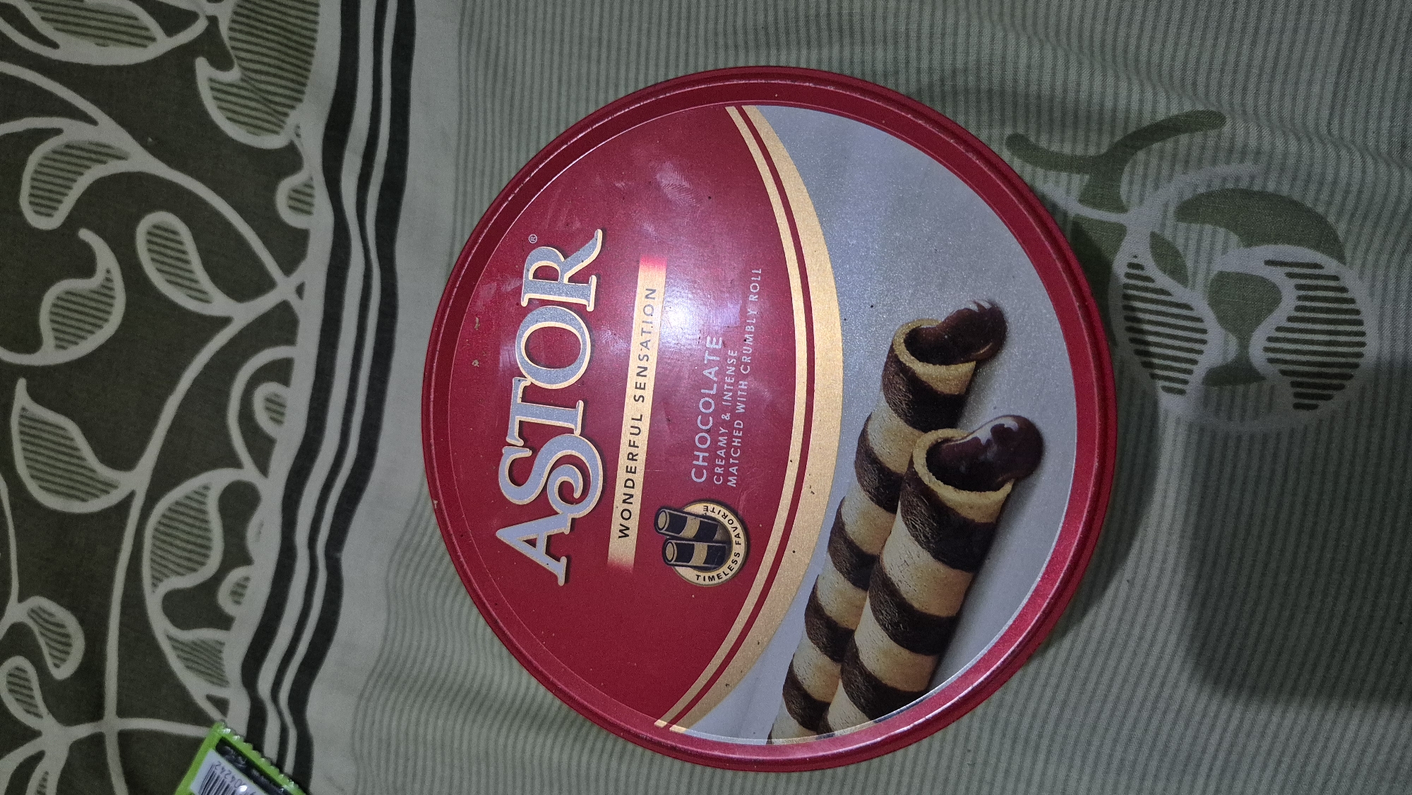 The image shows a round, red tin of Astor chocolate roll biscuits placed on a green and white striped patterned cloth. The tin has the brand name "ASTOR" prominently displayed in silver lettering at the top, with the tagline "WONDERFUL SENSATION" below it. A circular emblem with two chocolate rolls inside and the words "TIMELESS FAVORITE" is visible to the left of the text "CHOCOLATE CREAMY & INTENSE MATCHED WITH CRUMBLY ROLL". Two chocolate roll biscuits, partially broken to reveal their chocolate filling, are depicted on a gray surface within the tin's design. The background of the image is the patterned cloth, which appears to be a bedsheet or tablecloth. There are no people or actions depicted in the image. The image is an overhead shot of the product. It's not possible to determine the location in the city of Karawang, Indonesia from the image itself.