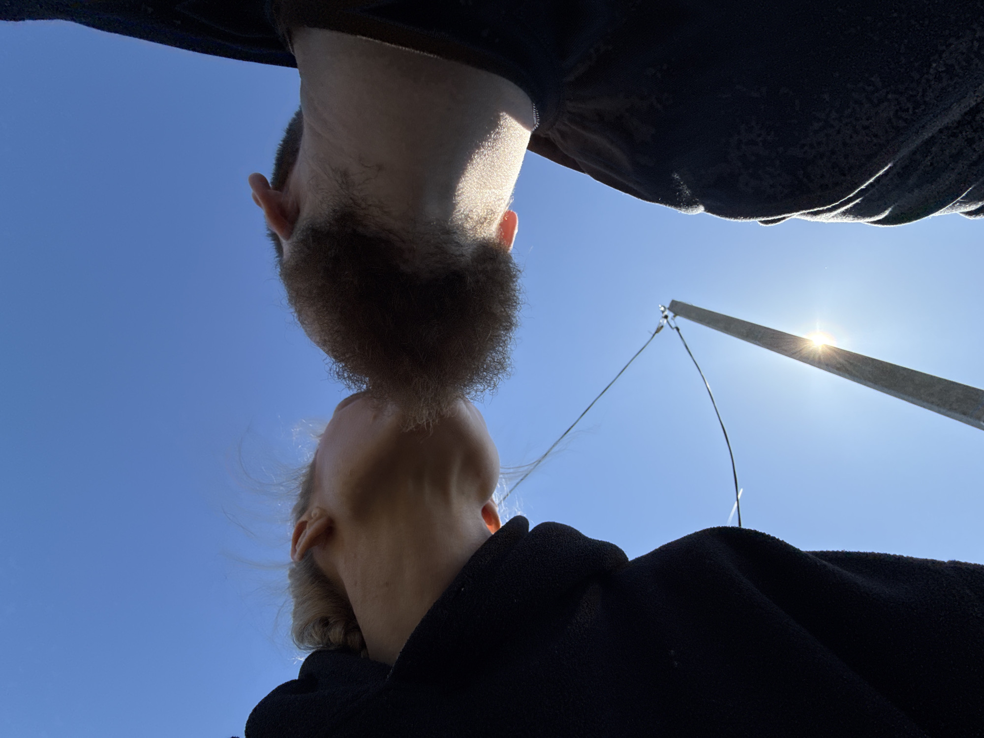 A low-angle shot, taken in daylight, showing two people close together against a clear blue sky. The person in the foreground is facing upwards, with their neck and chin visible. They have light skin and short, fair hair. They are wearing a dark-colored garment. Above them is a second person, also facing downwards, with their beard almost touching the first person's chin. This person has darker skin and a full beard. The perspective suggests the photo was taken looking straight up. In the background, a metal pole and wire are visible, catching the sunlight. The image does not obviously contain any information linking the location to Nizhnyaya Chermoda, Russia.