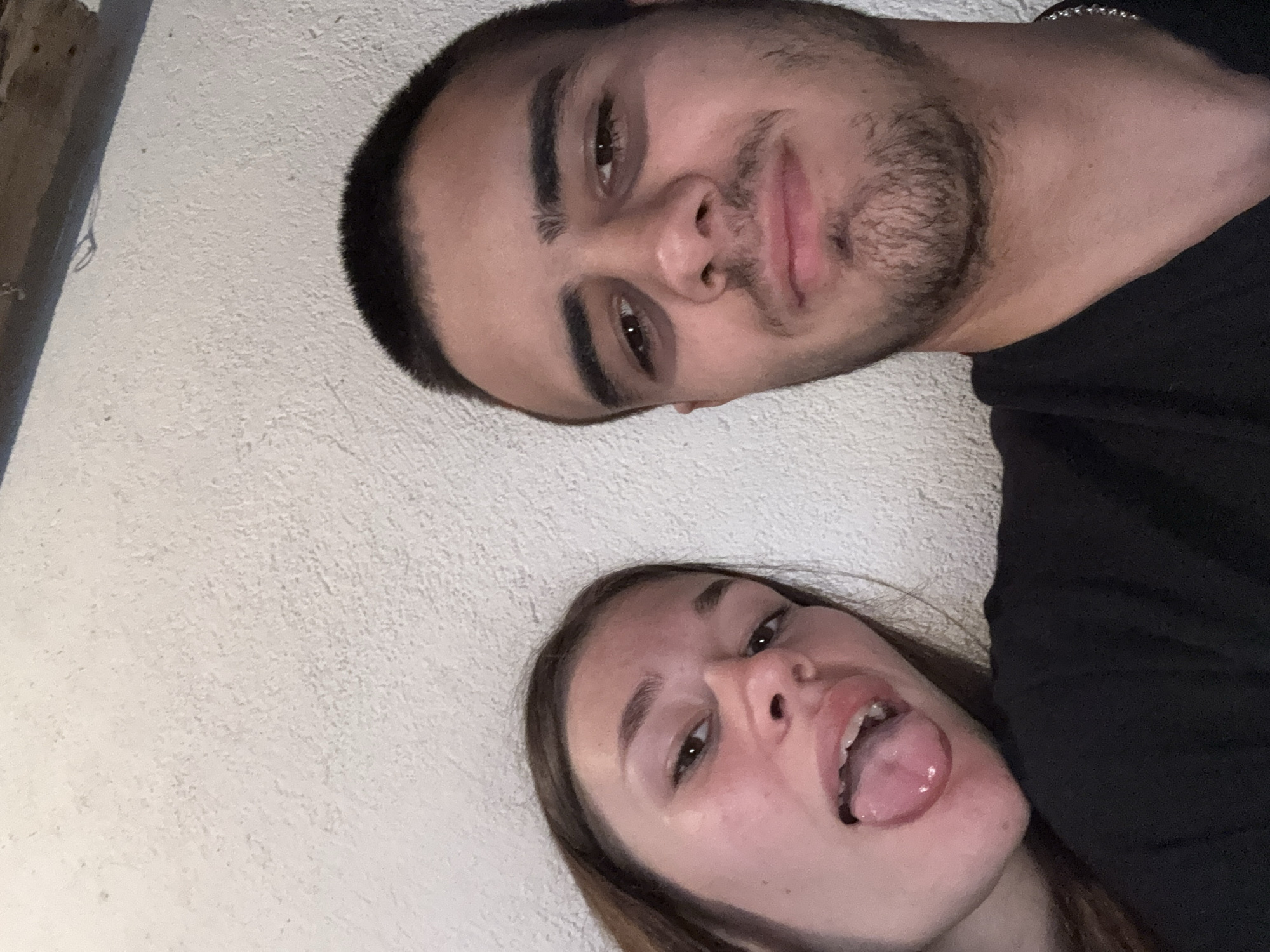A selfie shot shows two people posing for the camera. A man with short dark hair, a beard, and dark eyes is positioned at the top. Below him, a woman with long brown hair is sticking her tongue out. They are against a white wall. It is not possible to confirm that the picture was taken in Karlovo, Bulgaria.