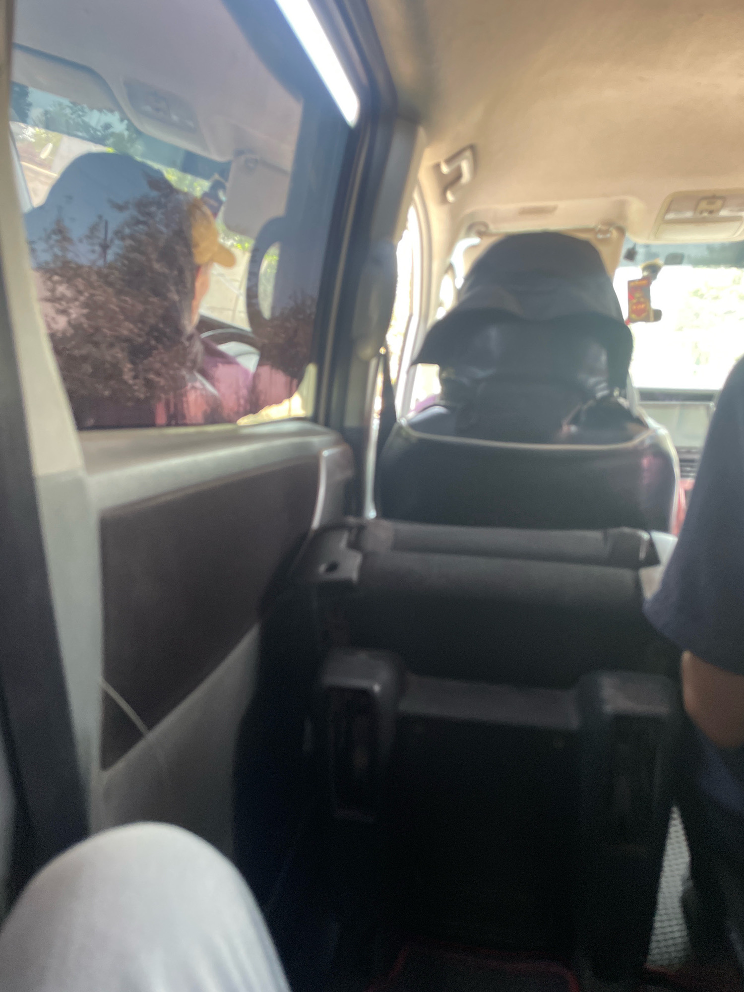 The image shows the interior of a vehicle, possibly a van or SUV, with multiple passengers. The perspective is from the rear passenger area, looking forward. In the foreground, a person's white leg and knee are visible. In the middle ground, the backs of several seats are seen, with one seat occupied by a person wearing a black head covering and a dark garment. To the left, a person wearing a yellow hat and a pinkish-purple top is visible through the side window, appearing to be in the driver's or front passenger seat. The exterior environment seen through the windows suggests a sunny day with trees and foliage. The location is identified as Pasarik, Tajikistan. The scene appears to be a typical commute or road trip within the city.