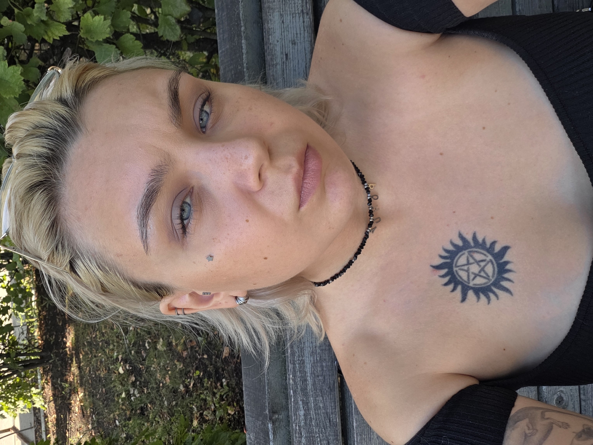 A close-up, chest-up, medium shot captures a young woman with blonde hair and blue eyes. She is outdoors, seated on a weathered wooden bench, with lush green foliage and dappled sunlight visible in the background. The woman wears a black off-the-shoulder top and a black choker necklace adorned with small letter charms. She has a star tattoo on her left cheek and a prominent pentagram tattoo on her chest. Her hair is windswept, framing her face. The image appears to be taken during daylight hours. The setting, given the context of Nizhny Novgorod, Russia, is likely a park or garden area within the city.
