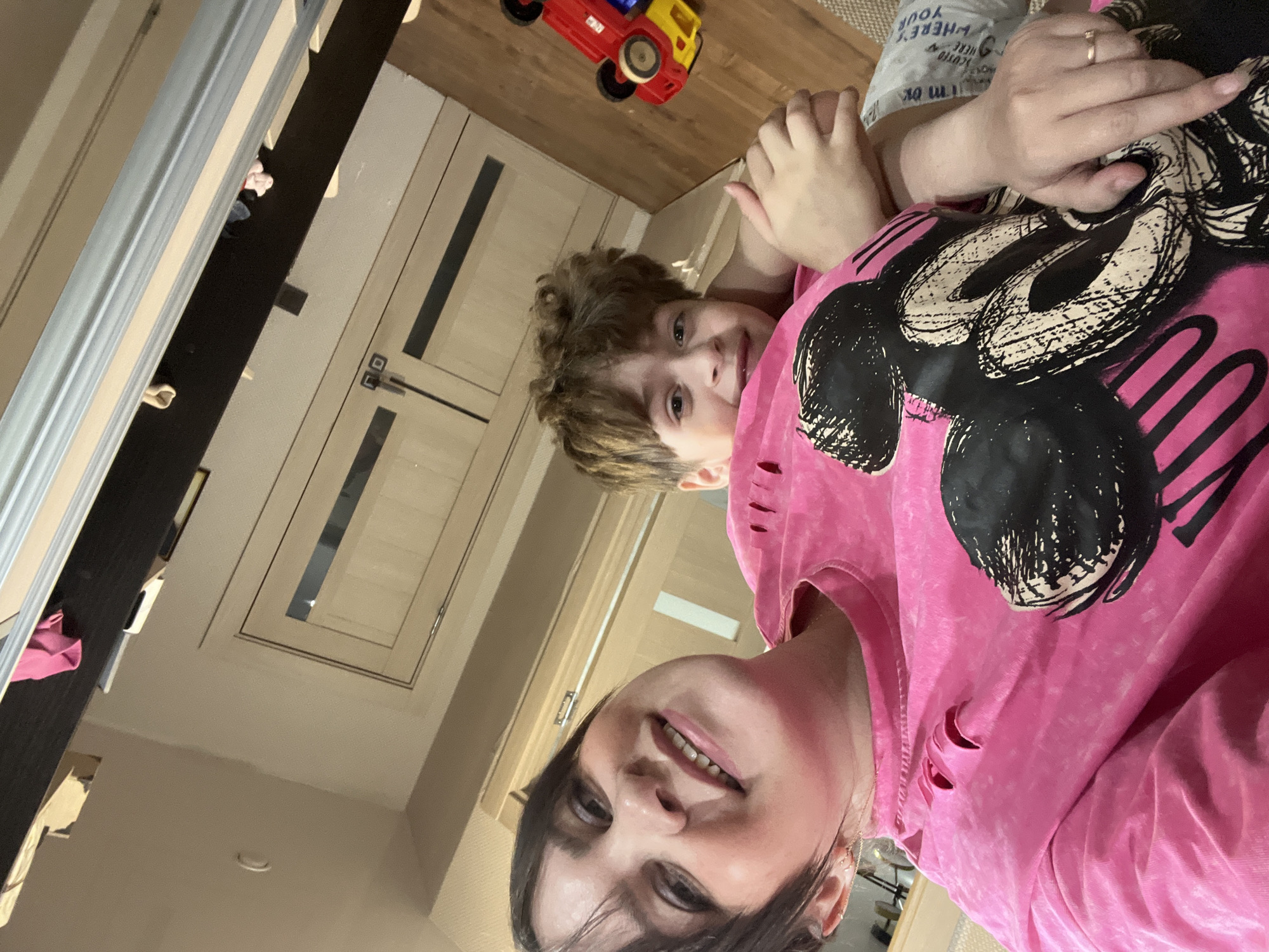 A selfie-style, indoor shot features a woman and a child. The woman, positioned in the foreground, is wearing a pink sweatshirt with a Mickey Mouse design. Her dark hair frames her face, and she is smiling. Behind her, a child with brown, curly hair is visible, also looking towards the camera. The background includes a wooden floor, a toy truck, and doors. The location is possibly in Noviy, Russia, as specified in the prompt. The image's perspective suggests it was taken from a high angle.