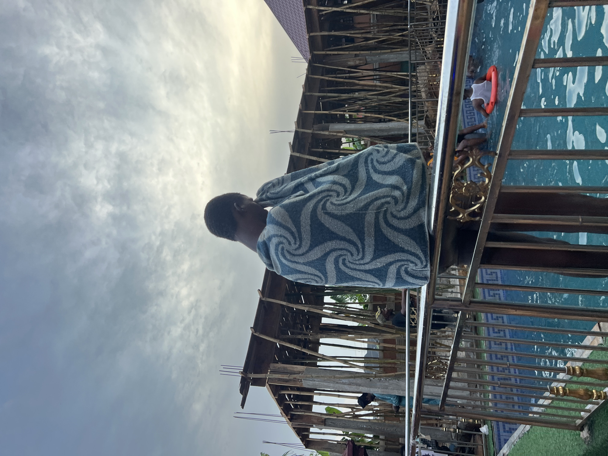 The image, when properly oriented, depicts an outdoor scene under a cloudy sky. In the foreground, viewed from behind, a person with short dark hair is partially visible, wrapped in a blue and white patterned towel or blanket. They appear to be looking towards the right side of the frame.
To the right of the foreground person, a metallic railing with ornate golden decorative elements separates the viewer's perspective from a swimming pool. In the mid-ground, a person wearing a white top is in the blue-tiled swimming pool, holding an orange life ring. The pool tiles feature a blue and white geometric pattern.
In the left mid-ground and background, a building under construction is visible. Its structure is supported by extensive scaffolding made of numerous wooden poles. Part of a reddish-brown roof can be seen on the building. Other indistinct figures are faintly visible near the construction site.
The sky is largely overcast with scattered clouds, and there's a brighter patch of light towards the top right, suggesting an overcast day rather than direct sunlight. The overall atmosphere appears calm. The location context provided is Nkpor, Nigeria, implying this scene takes place in that region. No visible text is present in the image.