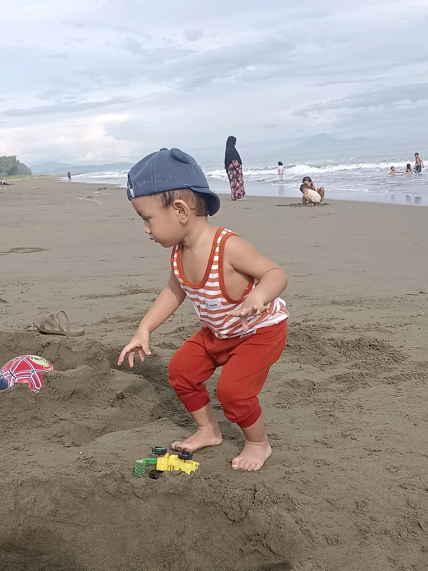 A young child wearing an orange and white striped tank top, orange pants, and a blue baseball cap with bear ears is playing in the sand on a beach. The child is crouched down, reaching towards a small yellow toy truck. A red, white, and blue ball and a pair of brown sandals are also visible in the sand near the child. In the background, the ocean with gentle waves meets a sandy shore under a cloudy sky. Several other people are scattered along the beach and in the water, including a woman in a black hijab and floral dress standing near the water's edge, and children playing in the surf. The location is identified as the city of Reudeuep, Indonesia.