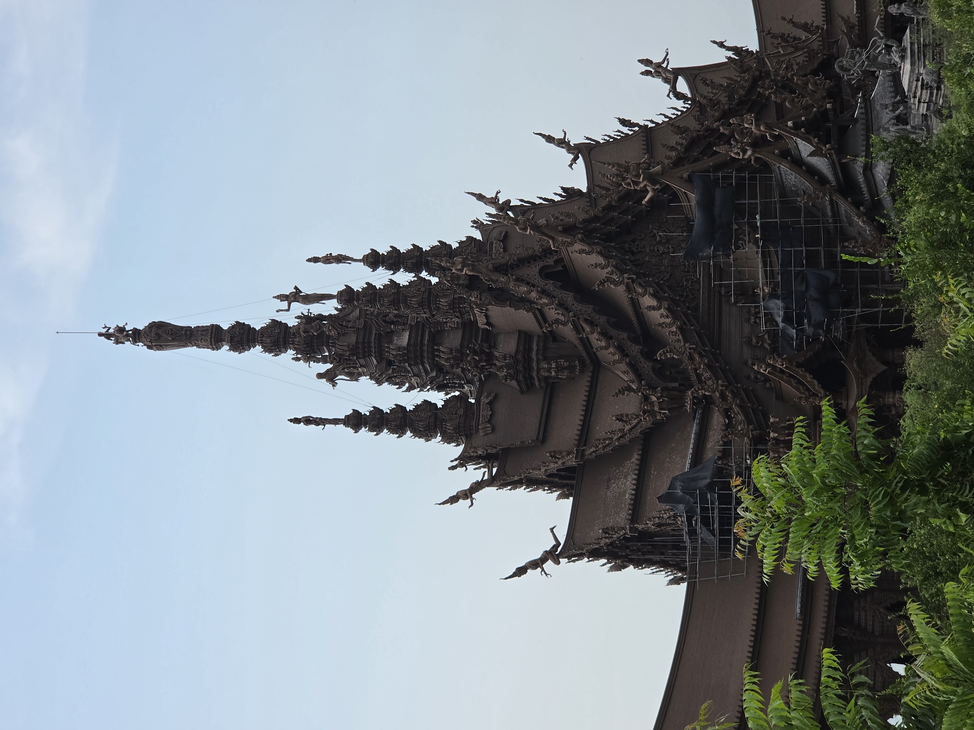 The image presents an upward view of the highly ornate and intricately carved wooden architecture of a temple or sanctuary, identified as the Sanctuary of Truth in Pattaya City, Thailand. The massive structure is crafted entirely from dark wood, showcasing numerous soaring spires, multi-tiered roofs, and elaborate gables.
The building is densely adorned with countless carved figures, depicting humanoids, deities, mythological creatures, and animals in dynamic poses along the rooflines, eaves, and ascending spires. These carvings are incredibly detailed, reflecting traditional Thai artistic and architectural styles. The uppermost spire, a prominent central feature, reaches towards the sky, topped with a finial.
The scene is set against a clear, light blue sky, indicating bright daytime conditions and good weather. On the right side and lower right foreground, vibrant green foliage from trees partially frames the wooden structure, adding a natural contrast to the man-made marvel. In the lower right section of the building, a metal scaffolding structure, partially covered with black tarps, is visible, suggesting ongoing preservation, maintenance, or construction work on the facade. No discernible text appears in the image. The overall impression is one of breathtaking craftsmanship, cultural richness, and spiritual grandeur.