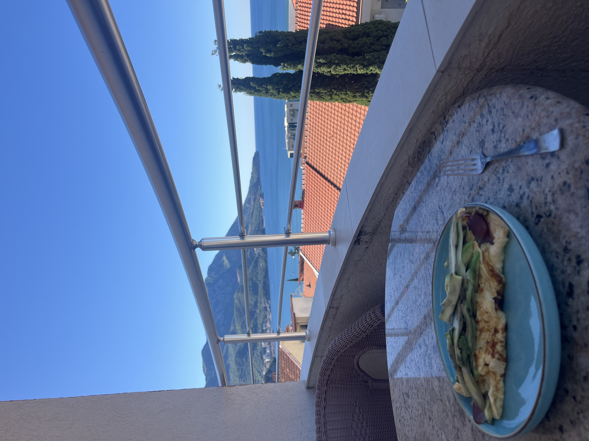 A plate of omelet with avocado and sausage sits on a marble-top table on a balcony overlooking a coastal city in Bečići, Montenegro. The omelet is on a blue plate with a brown rim. A fork rests next to the plate. In the background, red-tiled roofs of buildings are visible, along with two tall cypress trees. Beyond the city, the blue Adriatic Sea stretches to the horizon, meeting a clear blue sky. Mountains rise in the distance. A wicker chair is partially visible on the left side of the frame. The balcony railing is made of silver metal.