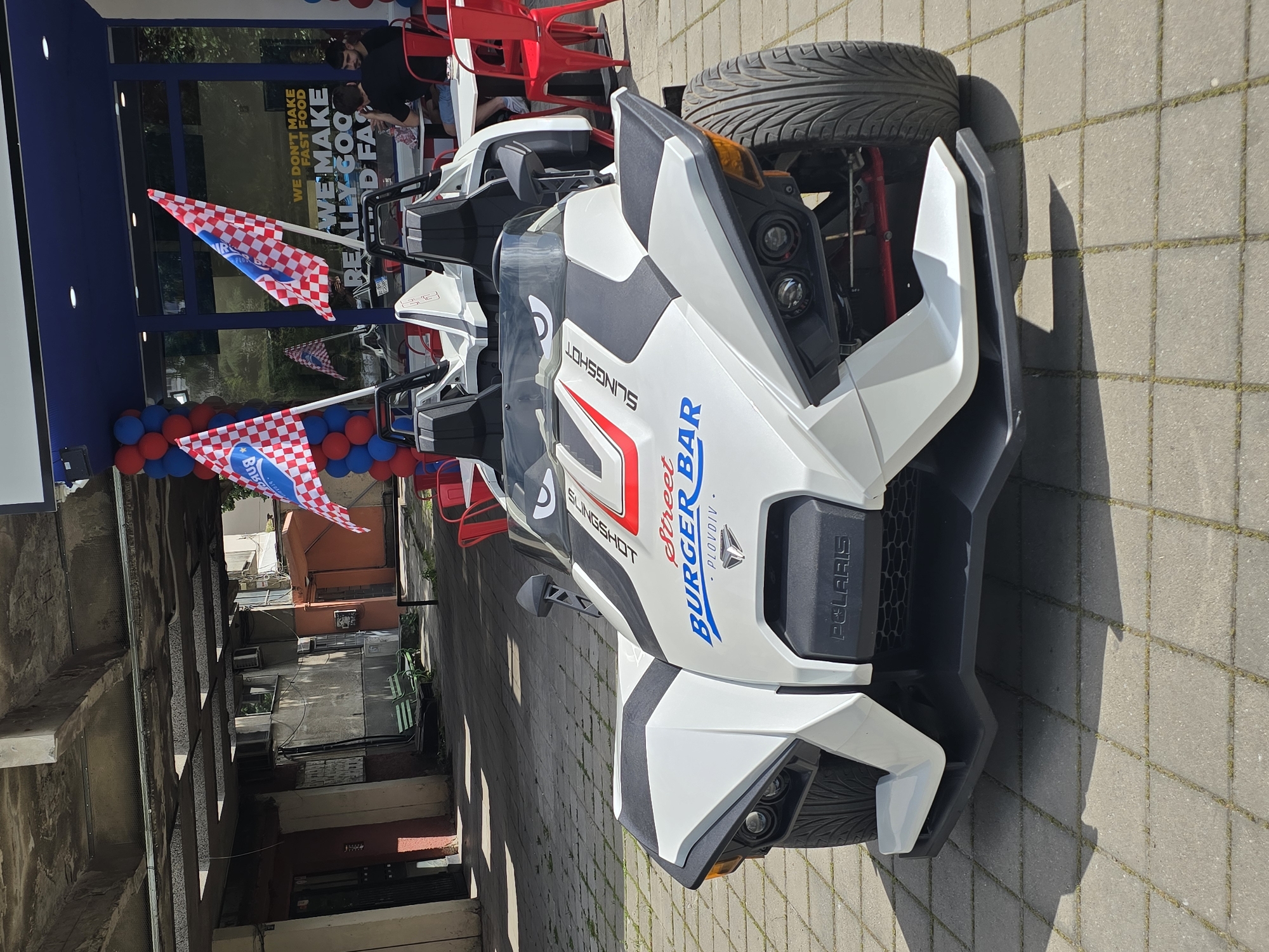 The image shows a white Polaris Slingshot three-wheeled vehicle parked in an urban environment, likely in front of a restaurant. The vehicle is adorned with "Street BURGER BAR" branding, suggesting a promotional tie-in with the establishment. Red and white checkered flags are attached to the Slingshot, adding a festive or competitive touch. In the background, a restaurant storefront is visible with balloons and outdoor seating. People are seated at the tables inside the restaurant. The scene appears to be outdoors on a sunny day, and the street is paved. The city is Unknown.