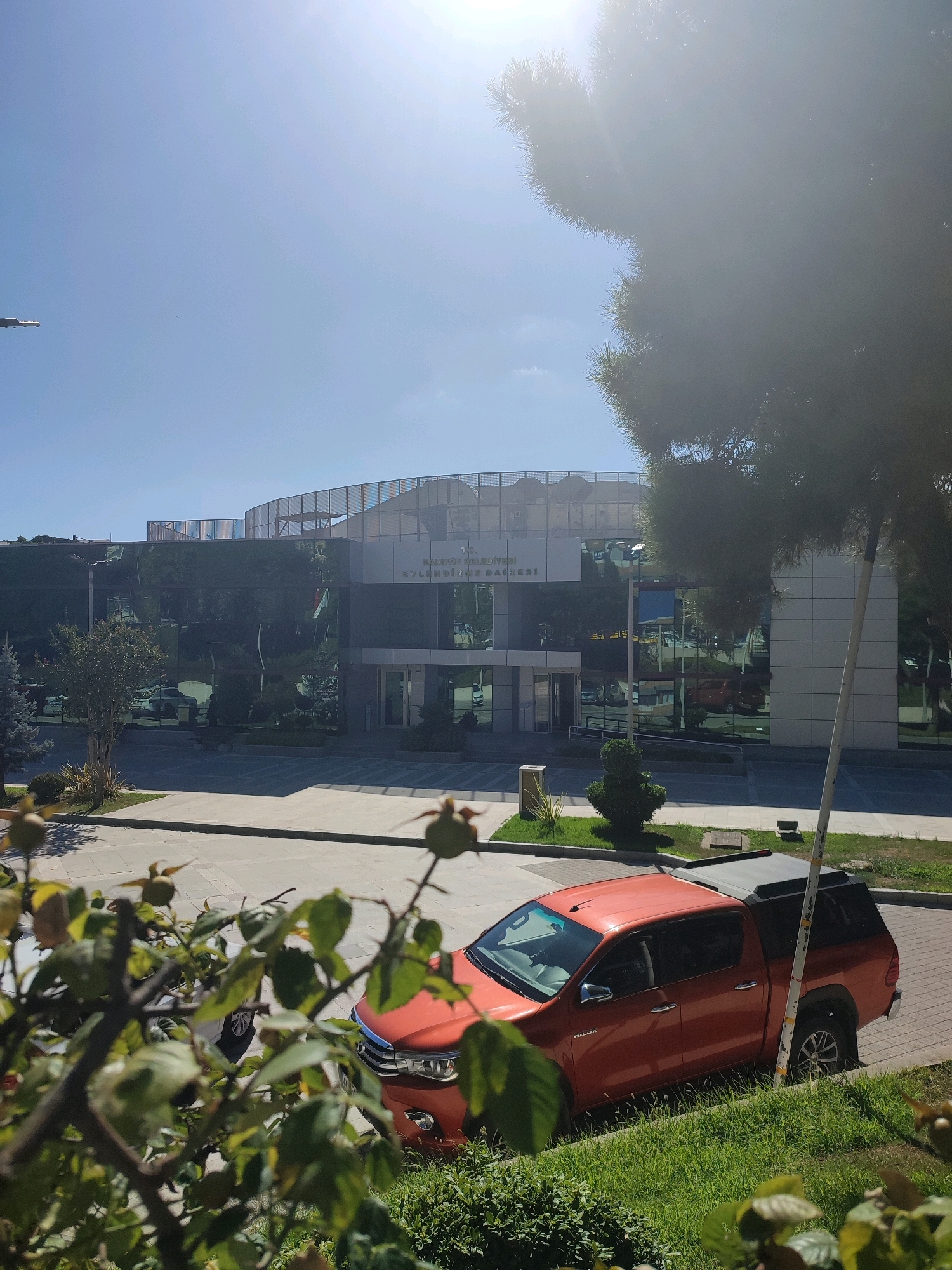 The image shows the exterior of the Kadıköy Municipality Building in Kadıköy, Istanbul, Turkey, on a sunny day. A modern glass-fronted building with a prominent signage displaying "T.C. KADIKÖY BELEDIYESI" and "Fen İşleri Daire Başkanlığı" is visible. The facade of the building is reflective, mirroring the surrounding trees and sky. In the foreground, out of focus, there are green plants and branches. To the right of the building, an orange pickup truck with a black canopy is parked on a paved area next to a grassy verge. The sky is clear and bright blue, with the sun creating a lens flare effect in the upper right corner.