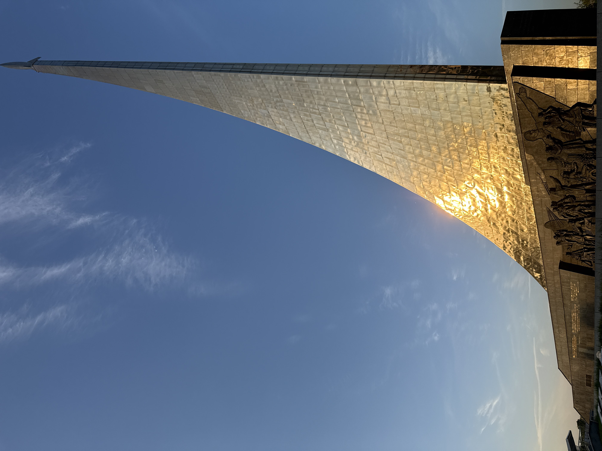 The image shows the Monument to the Conquerors of Space in Moscow, Russia. The monument is a tall, obelisk-shaped structure clad in reflective, metallic panels. The sun is reflecting off the panels, creating a bright, golden effect on the surface. To the right of the obelisk is a darker, rectangular structure adorned with bas-relief sculptures depicting figures and scenes. The sky is a clear, light blue with a few wispy clouds visible near the bottom left of the frame. The image is oriented vertically.