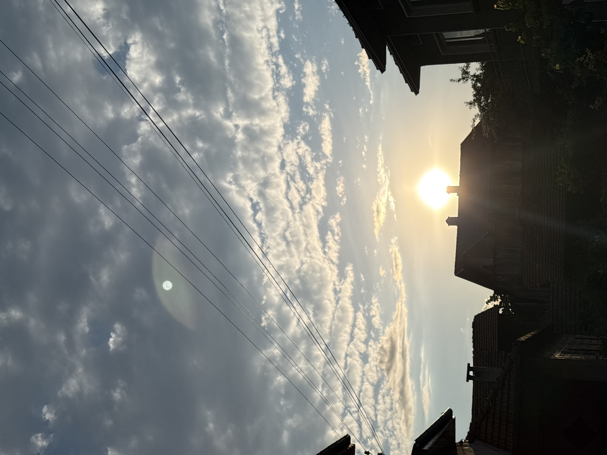 The image depicts a sunset over a village in Oreshak, Bulgaria. The sun is a bright, glowing orb near the horizon, casting a warm light that illuminates the sky and clouds. The sky is a mix of blue and grey, with scattered white clouds that appear to be illuminated from below by the setting sun. Several utility wires crisscross the sky, adding a man-made element to the natural scene. In the foreground, the silhouettes of houses with tiled roofs are visible. Some houses have chimneys. The overall impression is one of a peaceful evening in a rural setting.