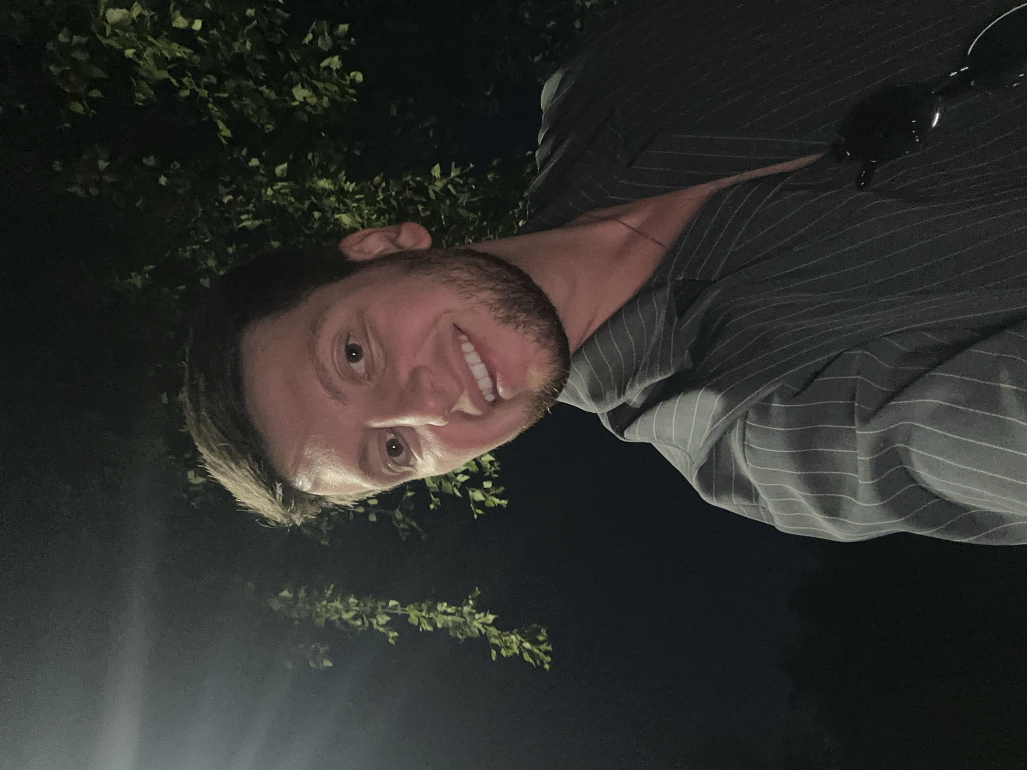 A man with short brown hair and a beard is smiling and looking at the camera. He is wearing a gray pinstripe shirt. Sunglasses are on his shirt. There are trees in the background. It is nighttime. The location is not discernible as Moscow, Russia.