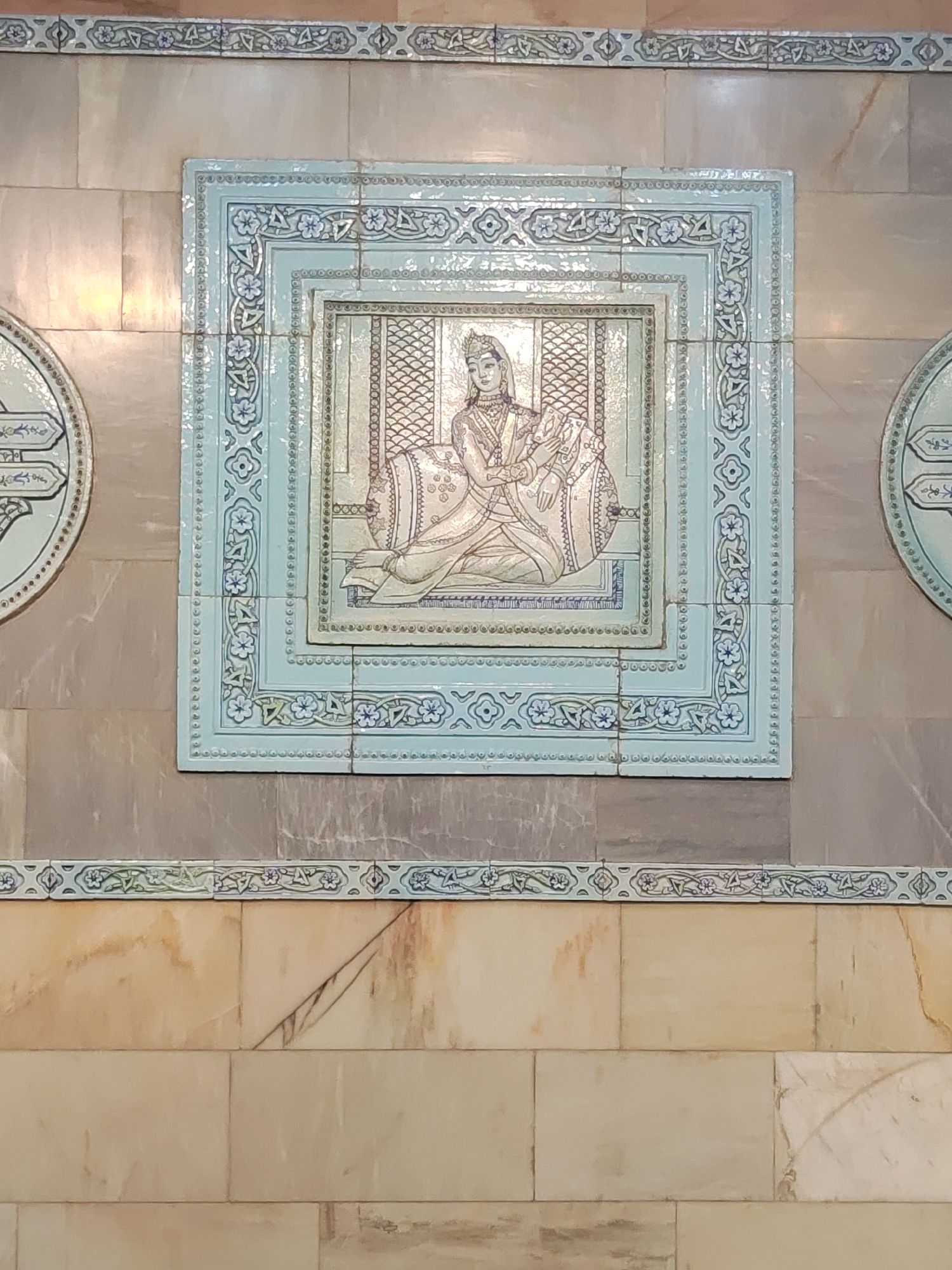 The image shows a close-up of decorative tiles on a wall. The central tile features a depiction of a person, likely a woman, seated on a cushion within an ornate frame. The figure is dressed in traditional attire, with a headdress and elaborate robes. The frame itself is adorned with intricate patterns and floral motifs in shades of blue and white. To the left and right of the central tile, partially visible, are circular decorative elements that appear to be part of the same tiled installation. The tiles are set against a background of larger, rectangular tiles with a marble-like pattern in muted tones of gray and beige. A decorative border with floral designs runs horizontally above and below the main tiled section. This tile work is a prominent feature of the Tashkent Metro, specifically found on the walls of the Amir Temur station, which is known for its rich ornamentation reflecting Uzbek history and culture.