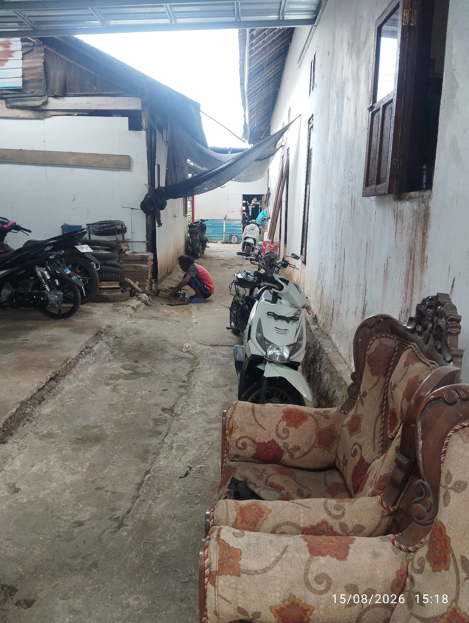 The image depicts an outdoor alleyway in Tegal Rejo, Indonesia, with a person engaged in activity. The alleyway is paved with concrete, showing signs of wear and tear with cracks and patches. To the left, several motorcycles are parked. A person is crouched on the ground near some stacked tires and what appear to be building materials, seemingly working on something. Further down the alley, more motorcycles are visible, along with a blue tarp. The right side of the alley is dominated by a white wall with several windows and a wooden-framed window that is open. In the foreground, a portion of an upholstered armchair with a floral pattern is visible, along with a timestamp in the bottom right corner: "15/08/2026 15:18". The overall environment suggests a functional, perhaps somewhat utilitarian, outdoor space.
