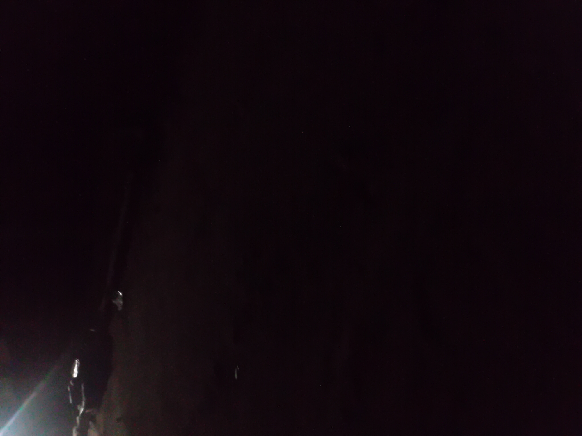 The image depicts a dark outdoor scene, likely at night, with limited visibility. A faint light source is visible in the upper left quadrant, casting a glow on a dark, amorphous object or surface. The rest of the image is predominantly black, suggesting a lack of ambient light. It is impossible to ascertain the location within Jauro Yinu, Nigeria, or any specific details about people, objects, or actions due to the poor lighting and low resolution of the image.
