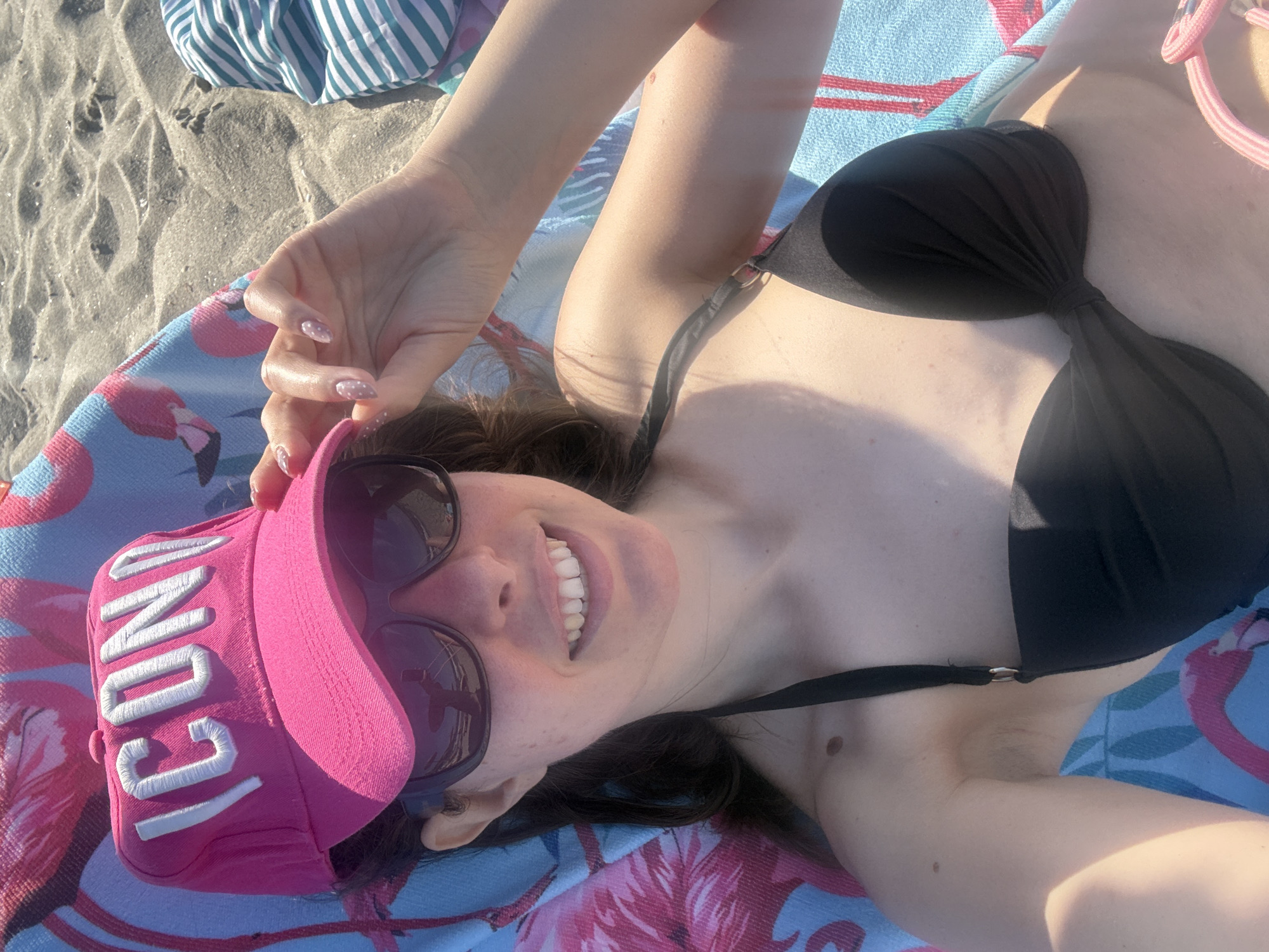 A person is lying on a beach towel on a sandy beach, viewed from a high angle. They are wearing a black bikini top and a pink baseball cap with white lettering that reads "ICON". Sunglasses with dark lenses are resting on their nose, partially obscuring their face. Their mouth is open in a smile, revealing white teeth. The beach towel has a pattern of pink flamingos. In the background, a striped towel and sand are visible. The lighting suggests it is daytime.