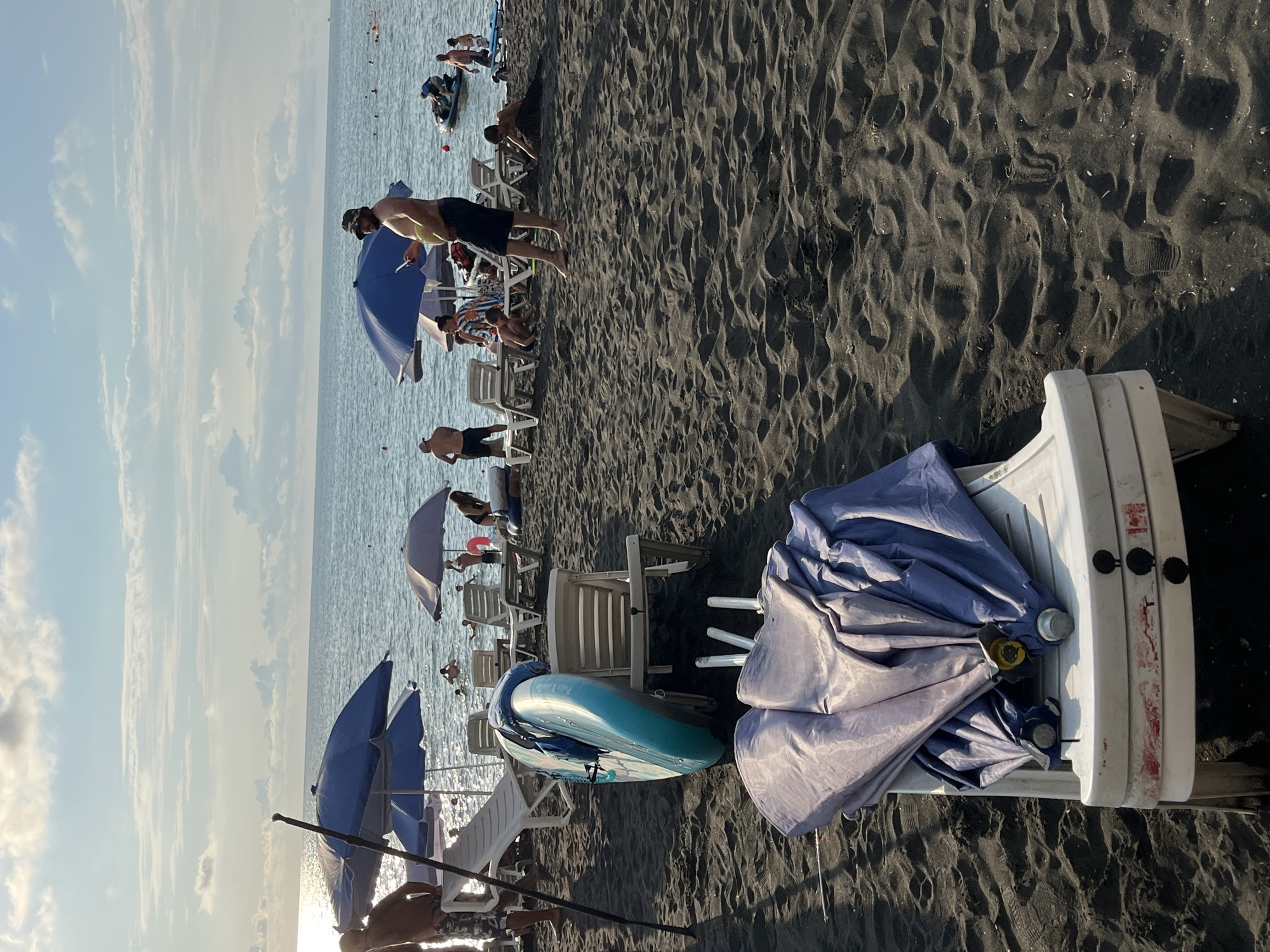 The image depicts a black sand beach in Ureki, Georgia, under a partly cloudy sky with the sun reflecting on the calm ocean. In the foreground, a white plastic beach chair is covered with folded purple and blue beach umbrellas, with their metal poles protruding. To the left, a turquoise stand-up paddleboard leans against a white lounge chair. Further back, several other white lounge chairs are scattered across the dark sand, some occupied by people, and several blue and grey beach umbrellas are open.
In the middle ground, the ocean is populated with swimmers and a few inflatable toys. A jet ski is visible in the distance. People are seen lounging on chairs, walking on the beach, and swimming in the sea. On the right side of the image, a person is partially buried in the sand, while another person watches from a short distance. The scene conveys a typical summer day at a coastal resort.
