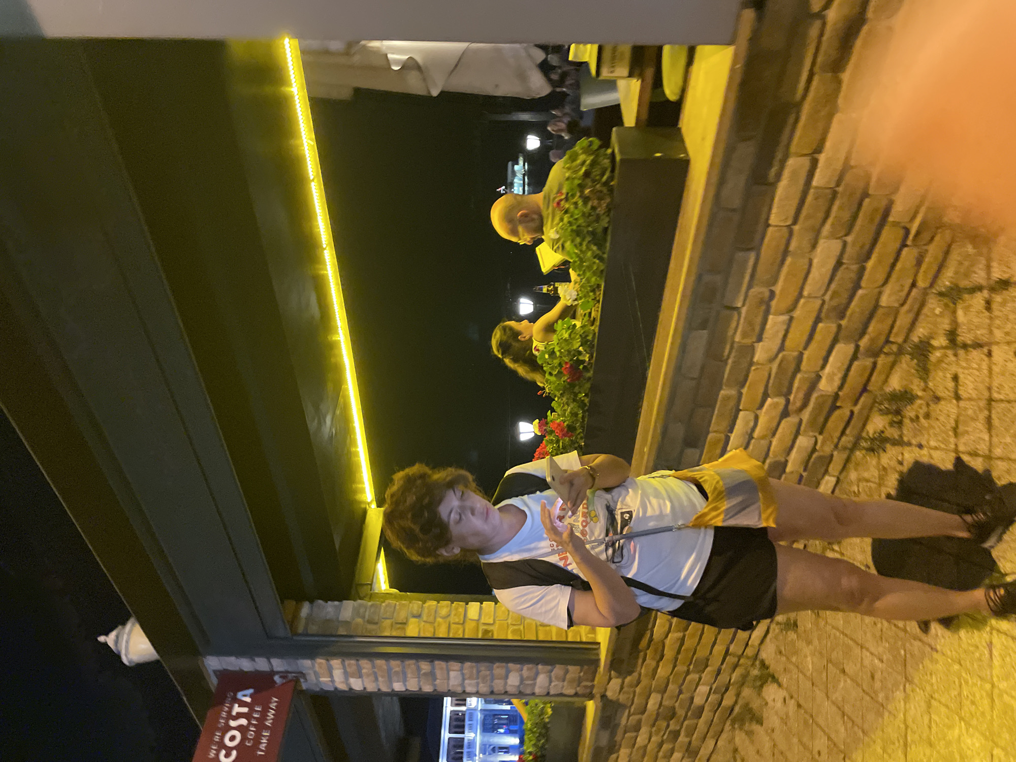 At night, a woman with curly brown hair stands in front of a brick wall, looking down at her phone. She wears a white t-shirt, black shorts, and a black backpack with a yellow and grey crossbody bag. Behind her, through an opening, two people are seated at a table outdoors. One person is a man with a bald head wearing glasses and a green shirt, facing away from the camera. The other is a woman with long dark hair facing the man. In the background, a building with lights is visible, and several streetlights illuminate the area. A red sign with "COSTA COFFEE TAKE AWAY" is mounted on the brick wall to the left of the woman. The scene appears to be an outdoor seating area of a cafe or restaurant in Kadıköy, Turkey.