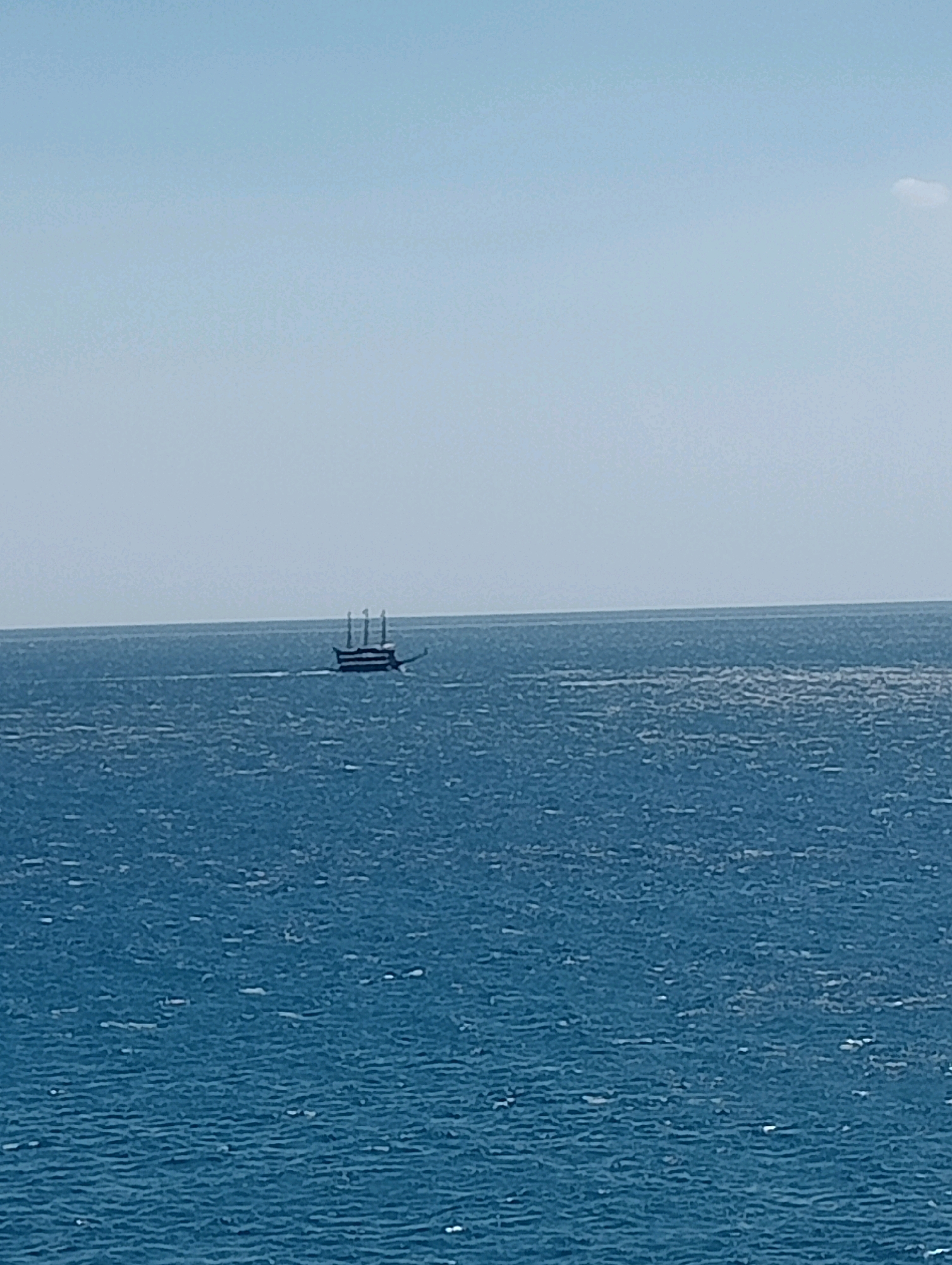 An image taken from a high vantage point shows a boat with three masts sailing on the ocean. The boat is dark in color and appears to be moving from left to right. The ocean is a deep blue with whitecaps, indicating choppy water. The sky is a pale blue, and there is a small cloud in the upper right corner. The horizon line is clearly visible, separating the sea from the sky. The image is likely taken in Antalya, Turkey, given the presence of boats and the clear blue sky.