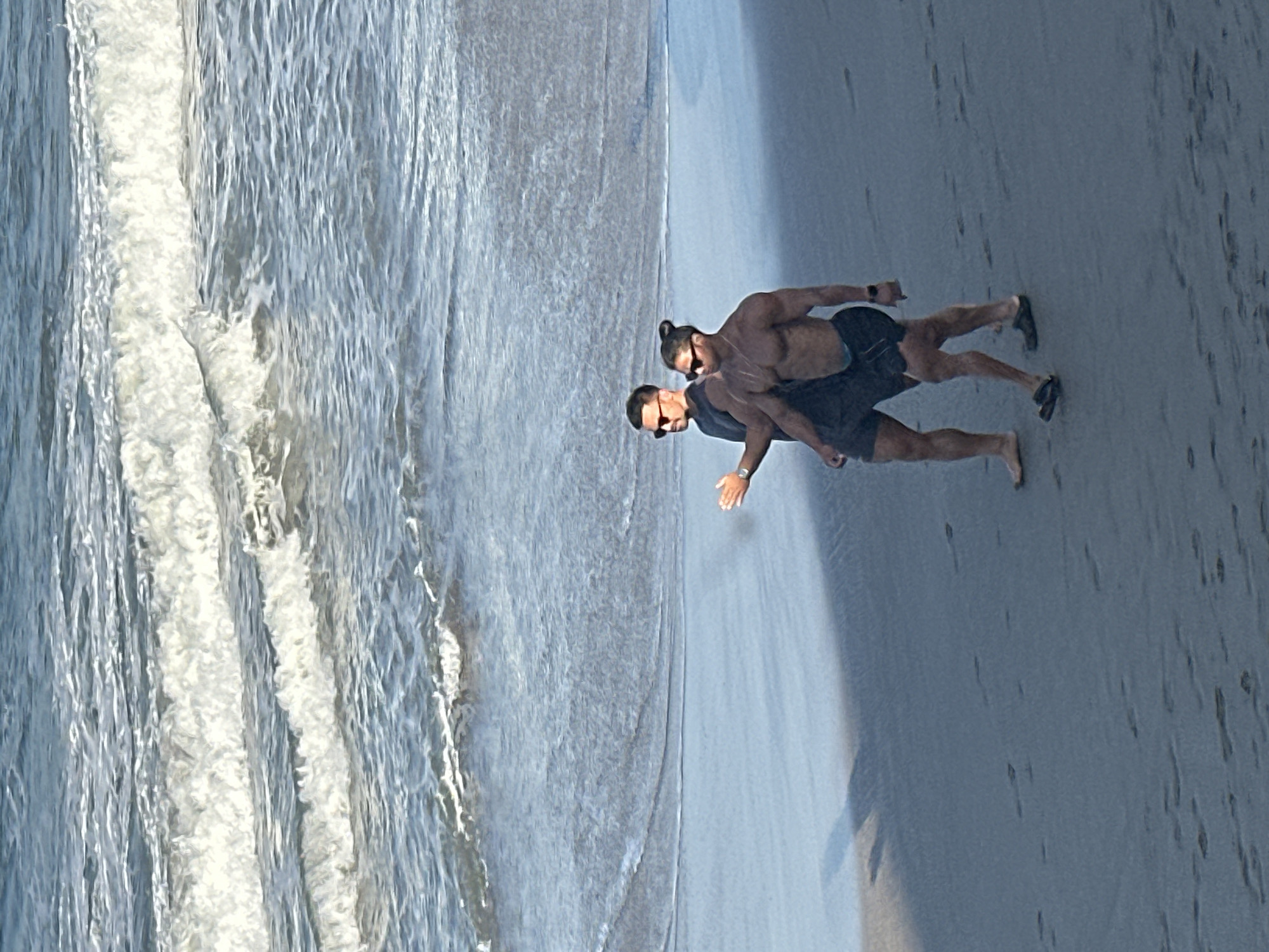 Two men are walking on a sandy beach near the ocean. The man in the foreground is shirtless with muscular build and wearing black shorts and sandals. The man behind him is wearing a dark t-shirt and shorts, and sunglasses. The ocean waves are visible in the background. The beach is mostly dark sand with some footprints. The scene appears to be in North Kuta, Indonesia.