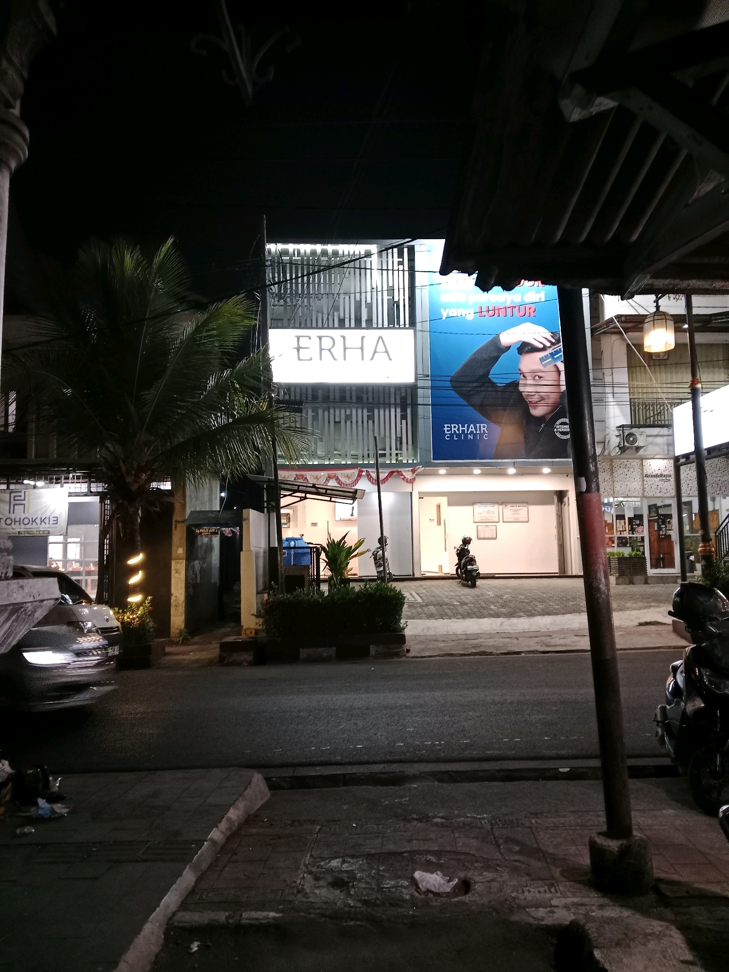 The image depicts a street scene at night in Cianjur, Indonesia, likely in a commercial area. A large, brightly lit building with "ERHA" signage is prominently featured. A billboard above the "ERHA" entrance showcases a man with a comb, advertising "ERHA CLINIC". To the left of the "ERHA" building, another establishment with "TOHOKKIE" signage is partially visible. A palm tree stands in front of the "ERHA" building. Several vehicles are present: a silver car with its headlights on is parked on the left, and a scooter is parked in front of the "ERHA" clinic. A motorcycle is parked on the right side of the image. The street is paved, and a sidewalk runs alongside it, with some debris visible on the sidewalk. A lamppost with a lit fixture is on the right side of the frame. The overall impression is a well-lit commercial street at night.