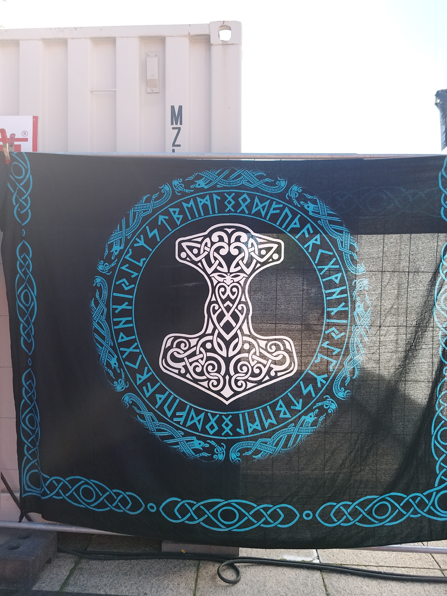 A black cloth banner with a Norse theme hangs in front of a white shipping container. The banner features a white, stylized representation of Mjolnir, Thor's hammer, at its center. Surrounding the hammer is a circle of turquoise Norse runes and knotwork designs. The edges of the banner are also decorated with turquoise Celtic-style knotwork. The banner appears to be hung in an outdoor urban setting, possibly in the city of Unknown, Unknown.