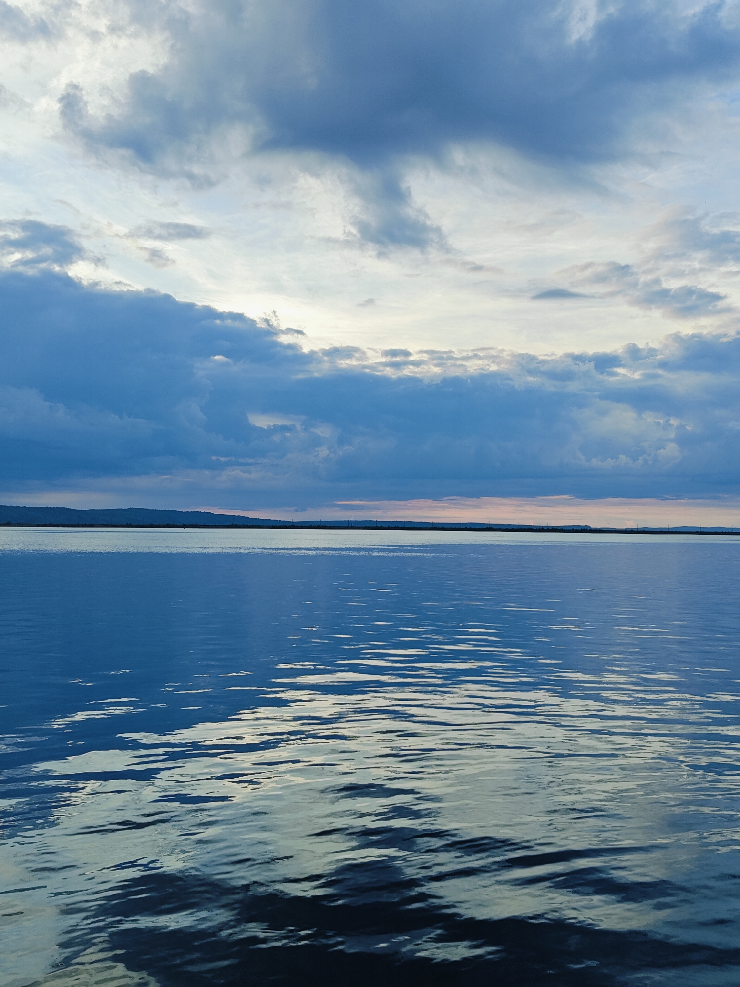 The image depicts a wide, calm body of water reflecting the sky, with a distant, low-lying shoreline. The sky is filled with dramatic, textured clouds in shades of blue, gray, and white, with hints of soft, warm light peeking through near the horizon, suggesting either sunrise or sunset. The water's surface is rippled, creating abstract patterns of light and shadow. The shoreline appears as a dark, silhouetted band against the lighter sky, with subtle undulations suggesting hills or a long stretch of land. There are no people, animals, or discernible structures visible in the image, creating a sense of vastness and natural tranquility. The overall impression is one of a serene, possibly remote, waterscape. This scene is likely located on the Kama River reservoir, near the city of Chaikovsky, Russia, a common feature of the region's landscape.