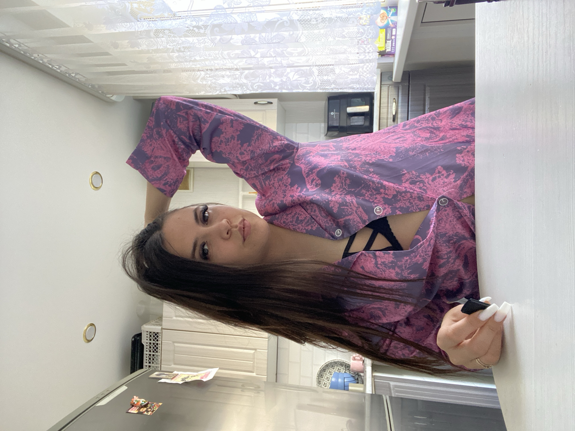 A woman with long, dark hair poses indoors in a kitchen setting. She wears a purple and pink patterned top revealing a black bra. Her manicured nails are visible as she touches a white surface, possibly a table or counter. Behind her, kitchen elements are visible, including cabinets, a microwave, and a refrigerator. The photo was taken indoors in Chaikovsky, Russia.