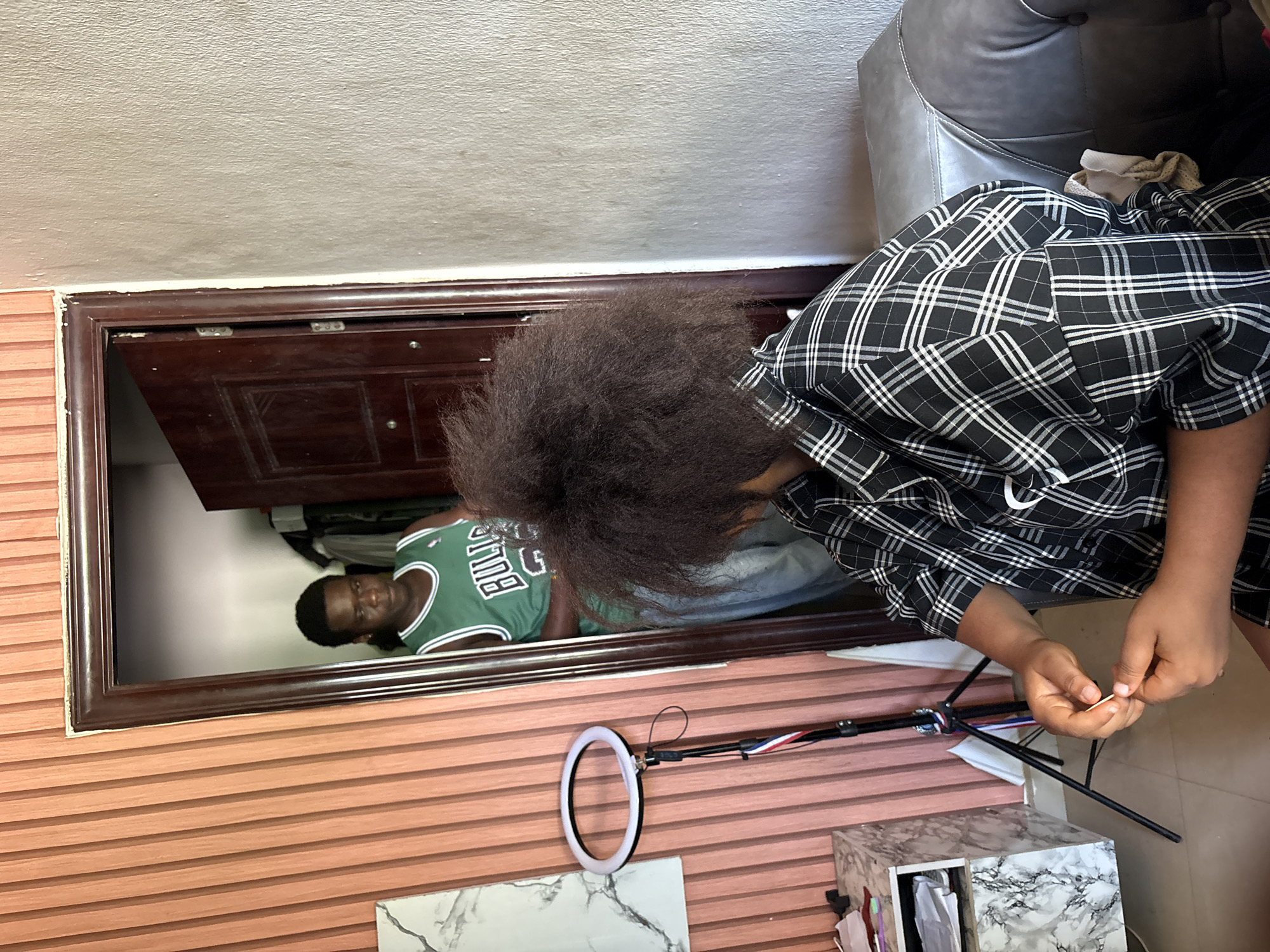 A room in Ikot Ekpene, Nigeria, contains a reflection of a person wearing a green Boston Celtics jersey in a mirror. A second person is seated in the foreground wearing a black and white plaid shirt. The person in the foreground is holding a toothpick. An open dark wood doorframe is visible behind the first person. A ring light is positioned near a marble-patterned cabinet. A grey leather couch is partially visible in the upper right corner of the frame. The walls appear to be painted white and a dark brown horizontally-patterned material.