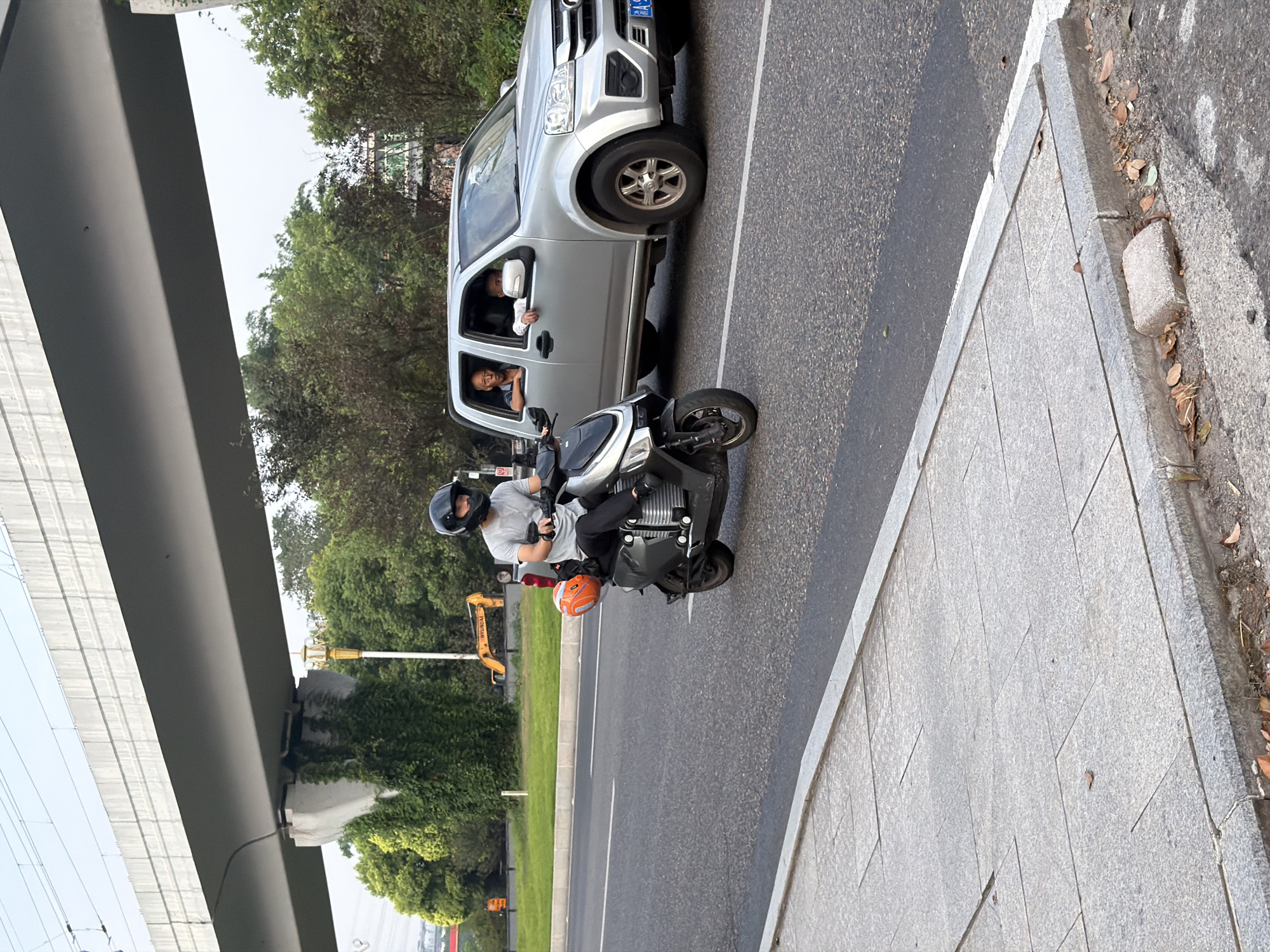 A man wearing a black helmet and a grey t-shirt rides a dark-colored electric scooter on a paved road in Jiangjin, China. He is wearing black pants and black socks. An orange helmet is attached to the rear of the scooter. To the right of the scooter, a silver pickup truck travels in the same direction. The truck has two occupants visible: an older man in the driver's seat looking forward, and a younger man in the passenger seat looking towards the camera. In the background, a concrete overpass spans the road, supported by columns covered in vines. Trees and foliage line the roadside, and a yellow excavator is visible behind a grassy embankment. The sky is overcast. The foreground shows a concrete sidewalk next to the road.