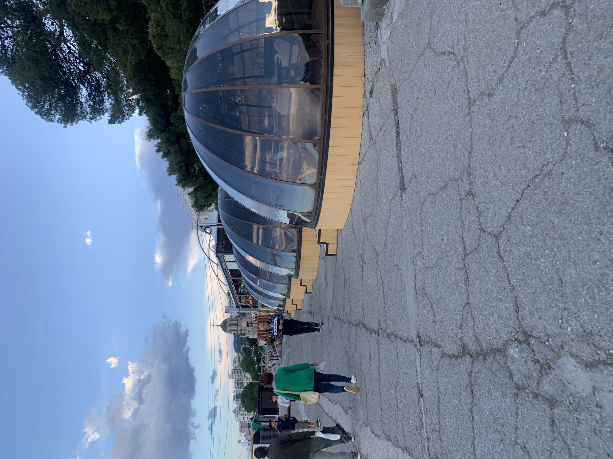 The image captures a street scene in Unknown, Unknown, featuring a row of dome-shaped structures with glass panels along the side of the road. The domes appear to be part of a restaurant or similar establishment. People are walking on the cracked sidewalk in front of the domes. The sky is visible with clouds. The street is cracked and weathered. In the background, city buildings are visible.