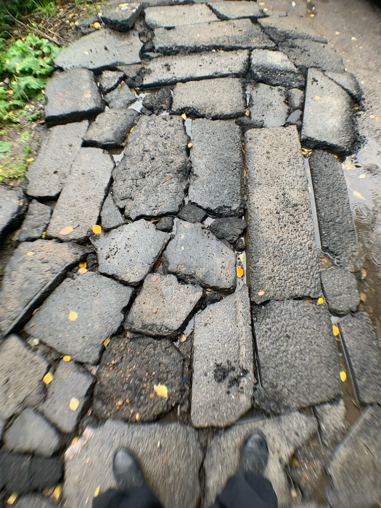 The image shows a close-up, downward-facing view of severely deteriorated asphalt pavement on a sidewalk in Arkhangelsk, Russia. The asphalt is broken into numerous irregular fragments, with large gaps and uneven surfaces. Puddles of water are present in the cracks and depressions, indicating recent rain. Scattered yellow autumn leaves are visible on the pavement. In the foreground, the legs and feet of an individual wearing dark boots and black trousers are visible, suggesting they are standing on or walking across the damaged sidewalk. The overall condition of the pavement indicates a significant lack of maintenance.