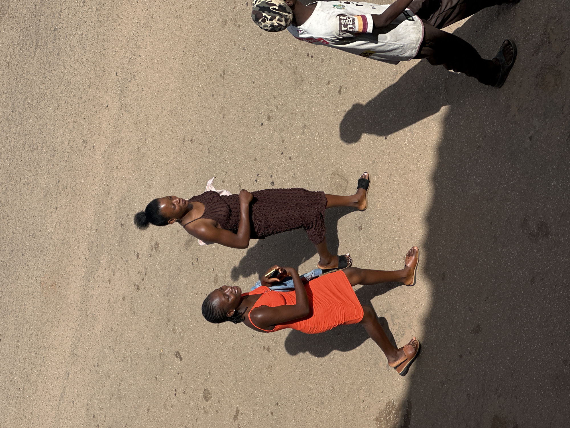 The image captures a street scene in Bukuru, Nigeria, from an elevated perspective. Three individuals are visible, walking across a paved road. On the left, a woman in a vibrant orange dress walks towards the right. She is carrying a blue object, possibly a phone or wallet, and wearing brown sandals. To her right, another woman in a textured brown dress is walking in the same direction, her hair tied up in a bun. She is also wearing sandals. Further to the right, a third person, partially obscured by shadow, is seen from the waist down, wearing dark pants, sandals, and a white shirt with a patterned head covering. The road surface is a mottled grey asphalt, showing signs of wear. Long shadows are cast across the pavement, indicating a bright, sunny day. The overall impression is a candid snapshot of everyday life on a Nigerian street.