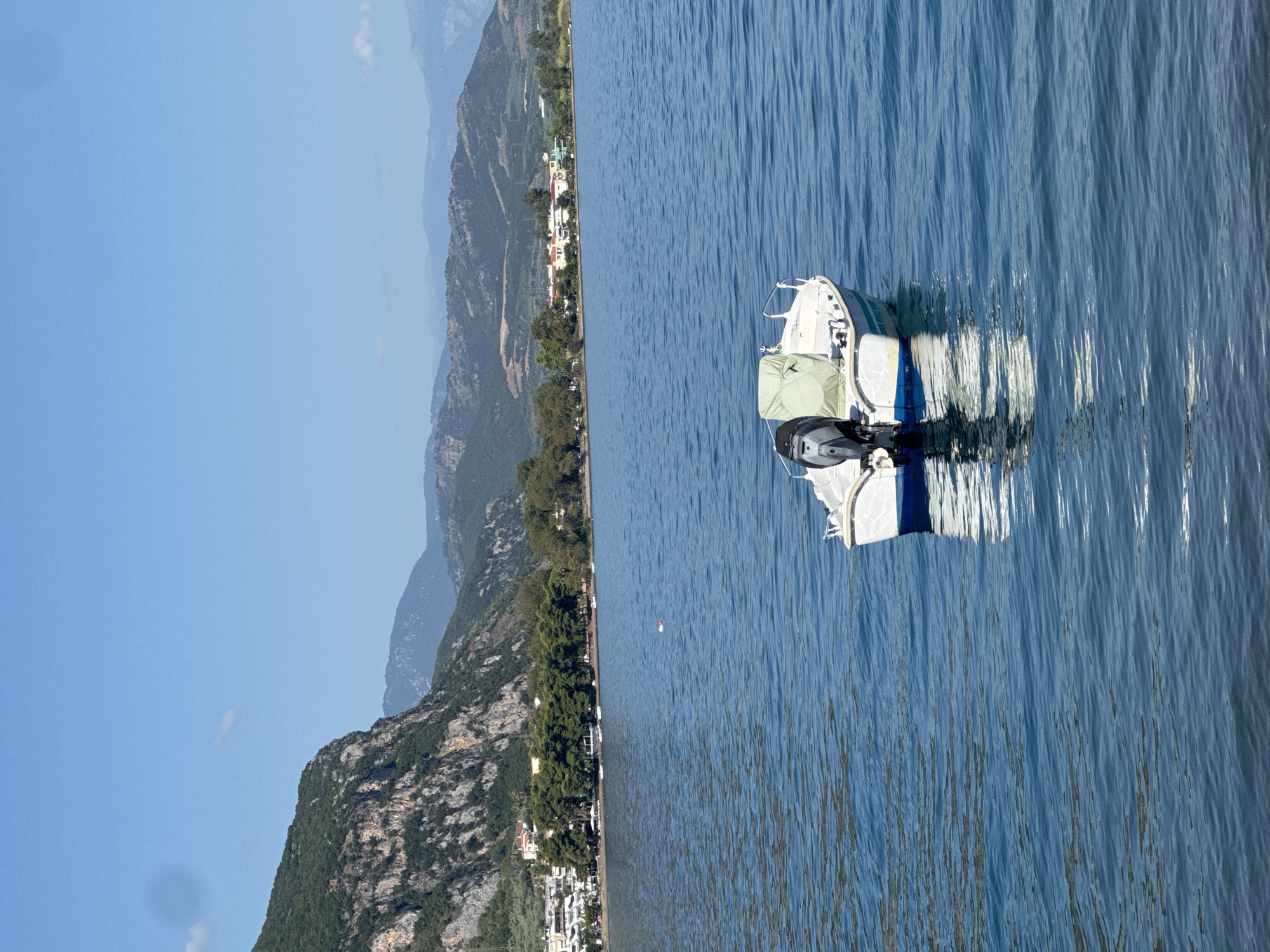 A small white boat with a blue stripe along its hull floats on a calm, blue body of water. The boat is powered by an outboard motor, and a light green tarp covers part of its interior. In the background, a coastline is visible, featuring a rugged, tree-covered mountain on the left and lower, rolling hills with scattered buildings and trees extending to the right. The sky is clear and blue with a few faint clouds. The scene appears to be within the Municipal Unit of Kamena Vourla, Greece, given the distinctive mountainous terrain bordering the water, characteristic of the region.