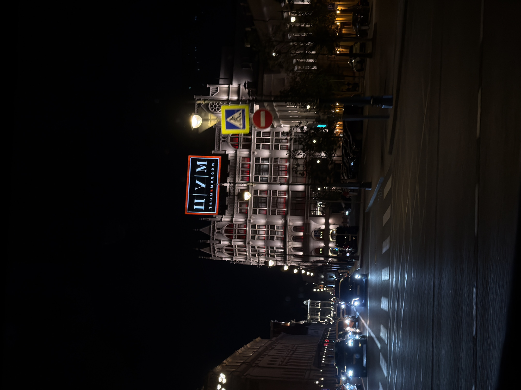 At night, the image shows a view of the city of Moscow, Russia. Triumfalnaya Square, with the Tsum department store building, is seen. The image is oriented vertically. Illuminated signs and road markings are visible. Cars are on the road.