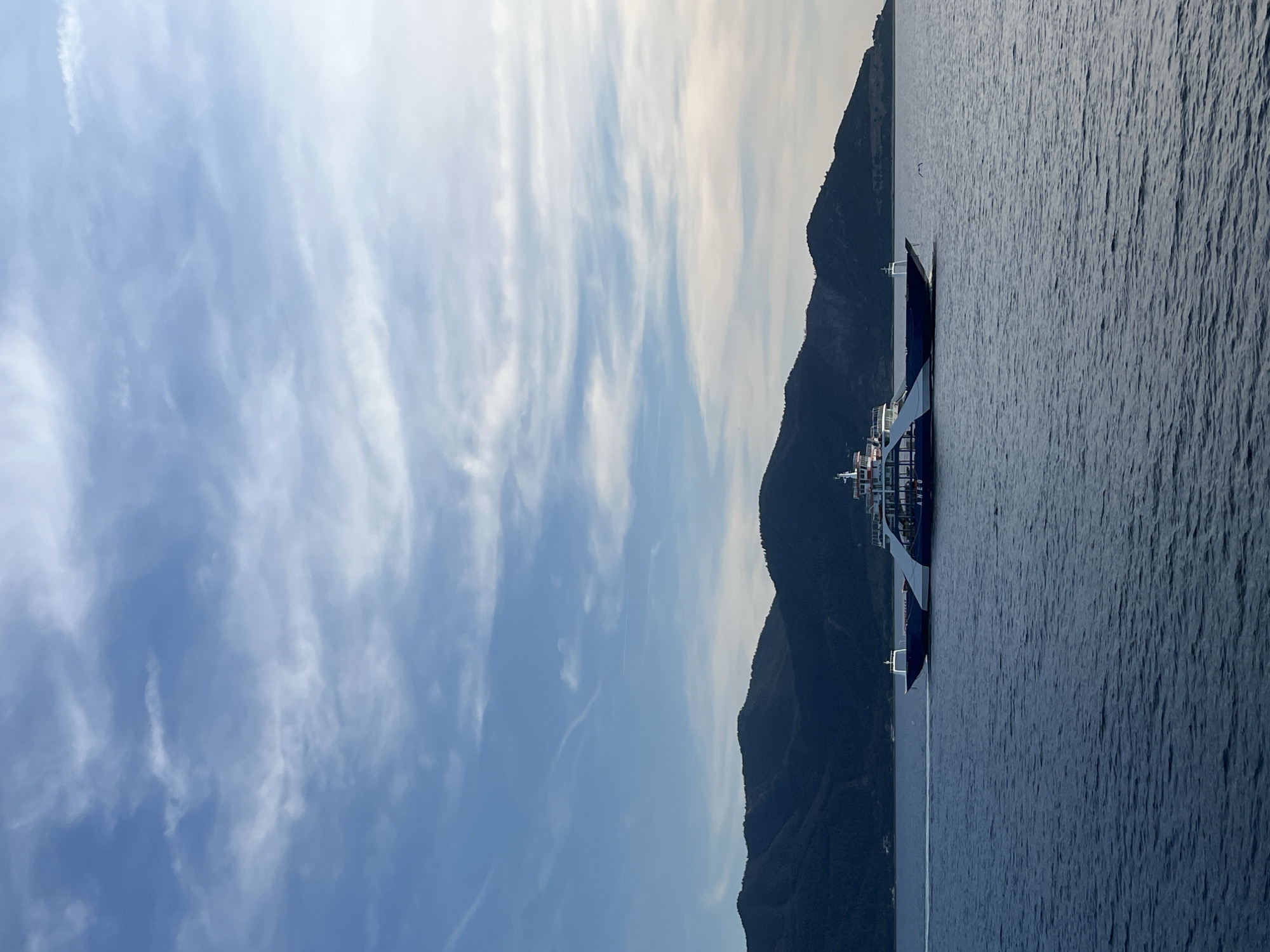 A ferry boat travels across a deep blue body of water, its hull dark blue and white, with the word "ANEO" visible on its side. The boat is moving from left to right, leaving a white wake behind it. In the background, dark green, mountainous terrain rises sharply from the water's edge, its peaks silhouetted against a pale blue sky filled with wispy, white clouds. The sky transitions to a lighter shade towards the horizon, indicating either early morning or late evening. The scene is peaceful and serene.