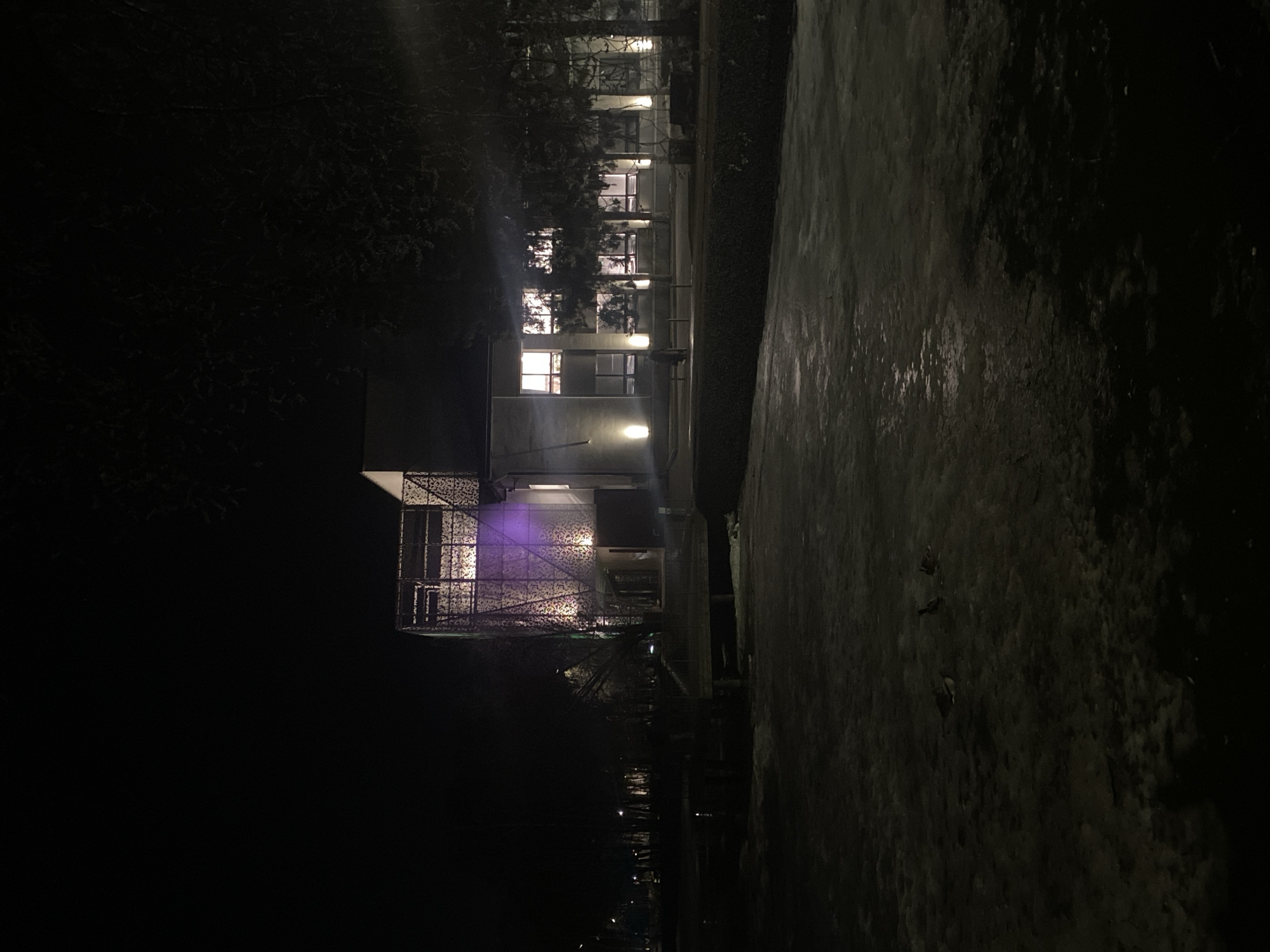 At night, a building with illuminated windows stands beside a dark, wet path. The structure features a decorative, possibly metal, lattice facade. Further down the path, there are indications of other lights or buildings in the distance. The city is Kazan, Russia.