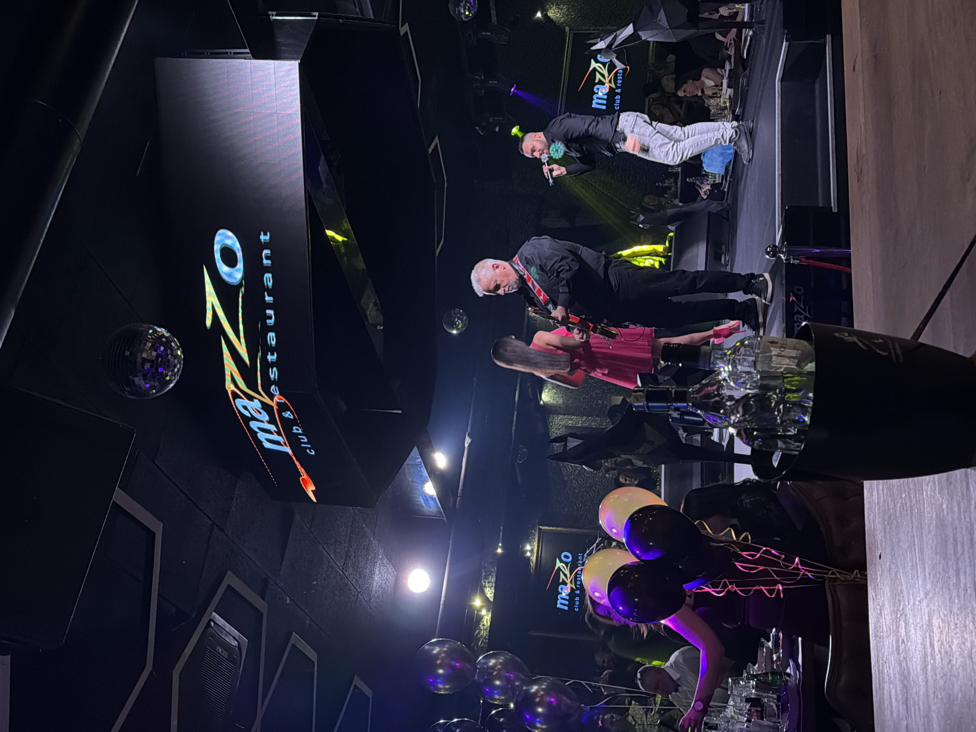 The image depicts an indoor scene at MaZZo club & restaurant in Sofia, Bulgaria, characterized by dim lighting and a live music performance.
In the immediate foreground, a dark wooden tabletop is visible, with an ice bucket containing multiple glass bottles, including one resembling a Johnnie Walker bottle and another partially visible, labeled "Polis." To the left, a bundle of balloons—two black, two yellow, and one purple—adorned with pink and gold ribbons, rises from a table where other drinks and glassware are present.
The central focus is a raised, dark-colored stage. On the stage, three individuals are engaged in a performance. A male with short grey hair and a black long-sleeved shirt with red decorative elements plays an electric guitar, positioned facing right. To his left, a female with long dark hair, wearing a bright fuchsia-pink dress, stands. To the right of the guitarist, a male vocalist with dark hair, dressed in a black shirt and light grey jeans, sings into a microphone. A green, spherical, textured accessory is visible on his left chest area. On the far right of the stage, a black, geometrically faceted sculpture, possibly depicting an animal, stands.
The background reveals numerous patrons seated at tables, observing the performance. The club's ceiling is dark, featuring decorative hexagonal and rectangular panels. Multiple disco balls are suspended from the ceiling. Several large illuminated screens display the venue's "MaZZo club & restaurant" logo in varying colors. Stage lighting includes directed spotlights and ambient colored lights, predominantly purple and green, illuminating the performers and surrounding areas. Visible air conditioning vents and industrial piping are present on the ceiling.