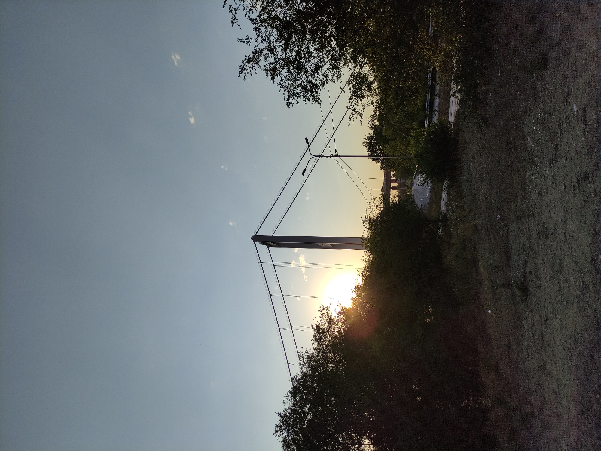 The image captures a sunset over a bridge structure in Semey, Kazakhstan. The sky is a clear, pale blue with a few wispy clouds. The sun is low on the horizon, casting a warm glow and creating lens flares as it shines through the trees. The main subject is a large suspension bridge, its towers and cables silhouetted against the sky. The foreground features overgrown vegetation, including bushes and trees, with a dirt and gravel path. In the distance, another bridge or overpass is visible, with vehicles on it. A streetlamp stands to the right of the main bridge structure, with power lines running between the bridge and the lamp. The overall impression is one of an industrial or transportation hub at the end of the day, with nature reclaiming parts of the landscape.