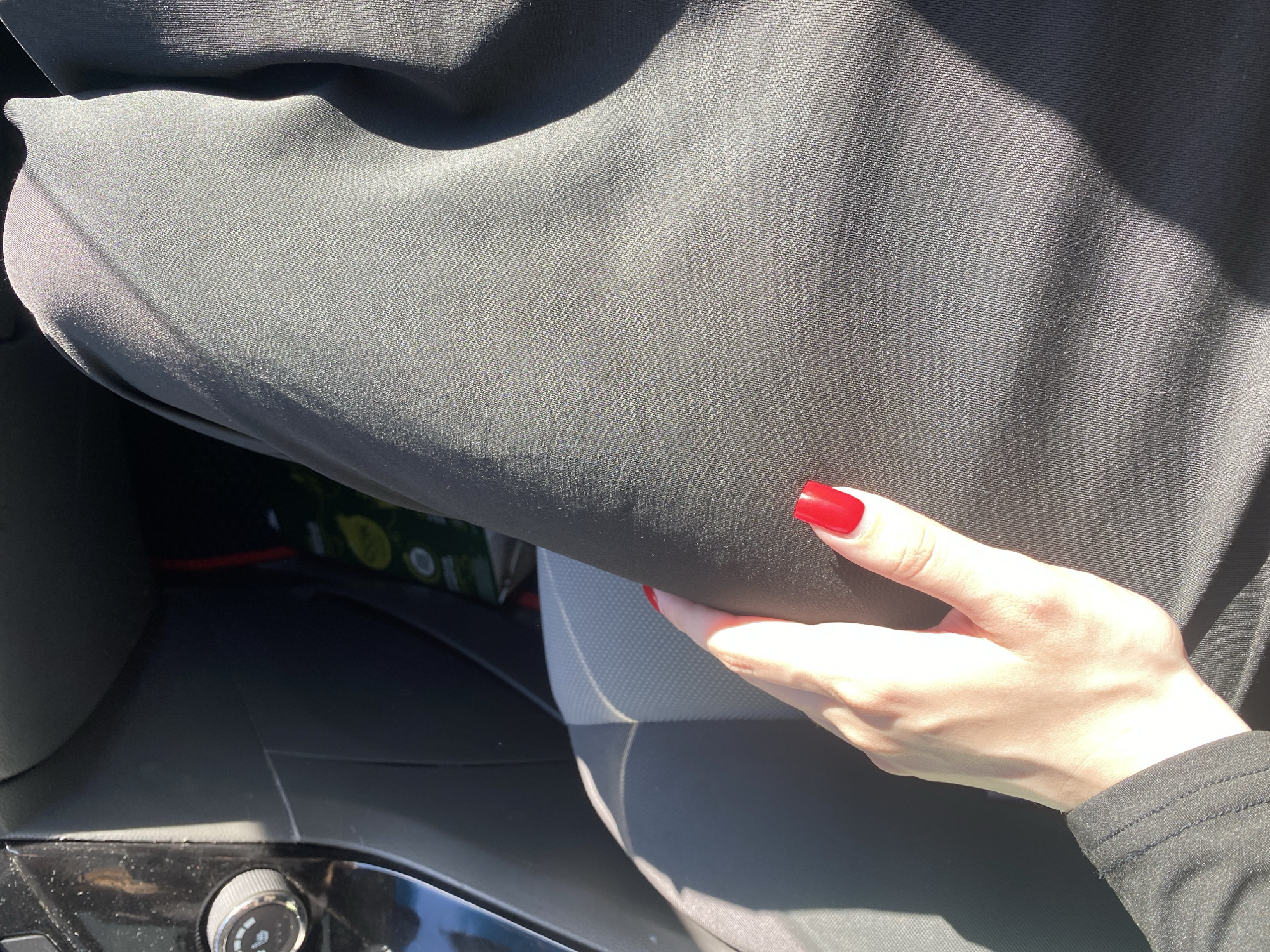 A hand with bright red, square-shaped fingernails is holding up a piece of black fabric. The fabric appears to be part of a car's interior, possibly a seat cover or upholstery, as a grey car seat is visible beneath it. The hand is illuminated by sunlight, casting shadows and highlighting the veins on the back of the hand. The person's skin is fair. Behind the fabric and seat, a glimpse of what might be a car's cargo area or floor is visible, with what looks like packaging for a product with a yellow and green design. The overall setting appears to be inside a car during the daytime, given the strong sunlight and shadows. The location is identified as Dyakovo, Russia. There is no visible text in the image. The image is a close-up shot, focusing on the hand and the fabric, suggesting an emphasis on texture and color contrast.