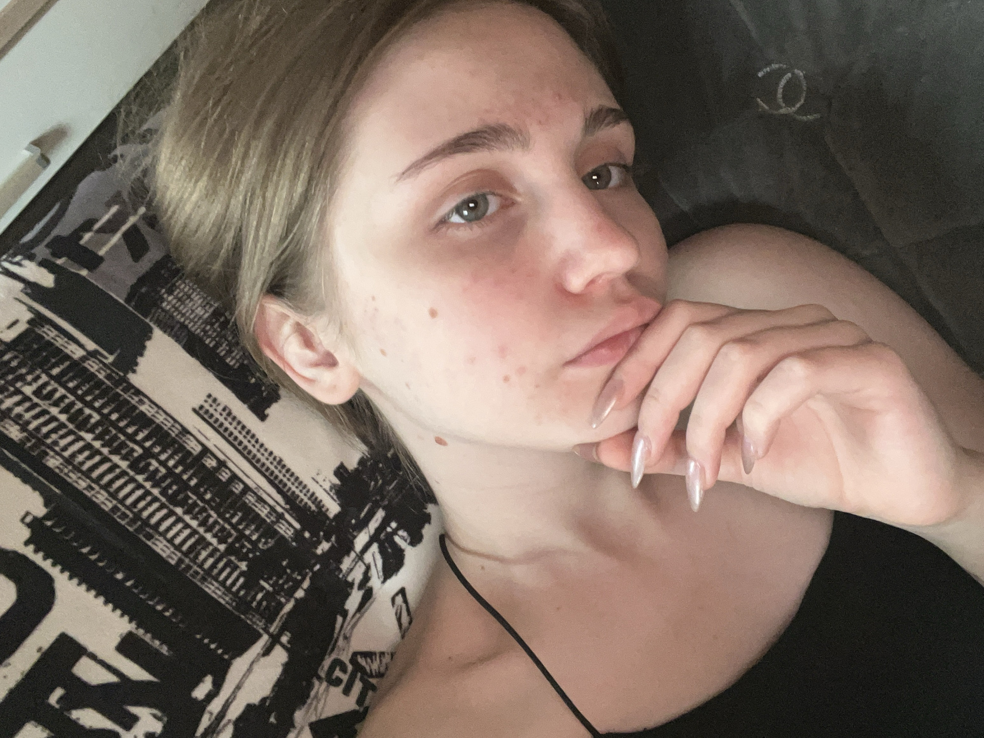 This is a close-up, high-angle selfie of a young woman with light brown hair and fair skin. She is lying on her side, with her head resting on a dark gray surface with a subtle circular logo. Her gaze is directed towards the camera, and her left hand is resting on her chin and mouth area. She is wearing a black, spaghetti-strap top. Her nails are long and appear to be painted with a pearlescent polish. The background consists of a black and white fabric with a repetitive pattern of what appears to be city buildings or abstract shapes. The lighting is soft and diffused, casting gentle shadows. The image does not contain any information that would allow for its location to be identified in the city of Ashkadar, Russia.