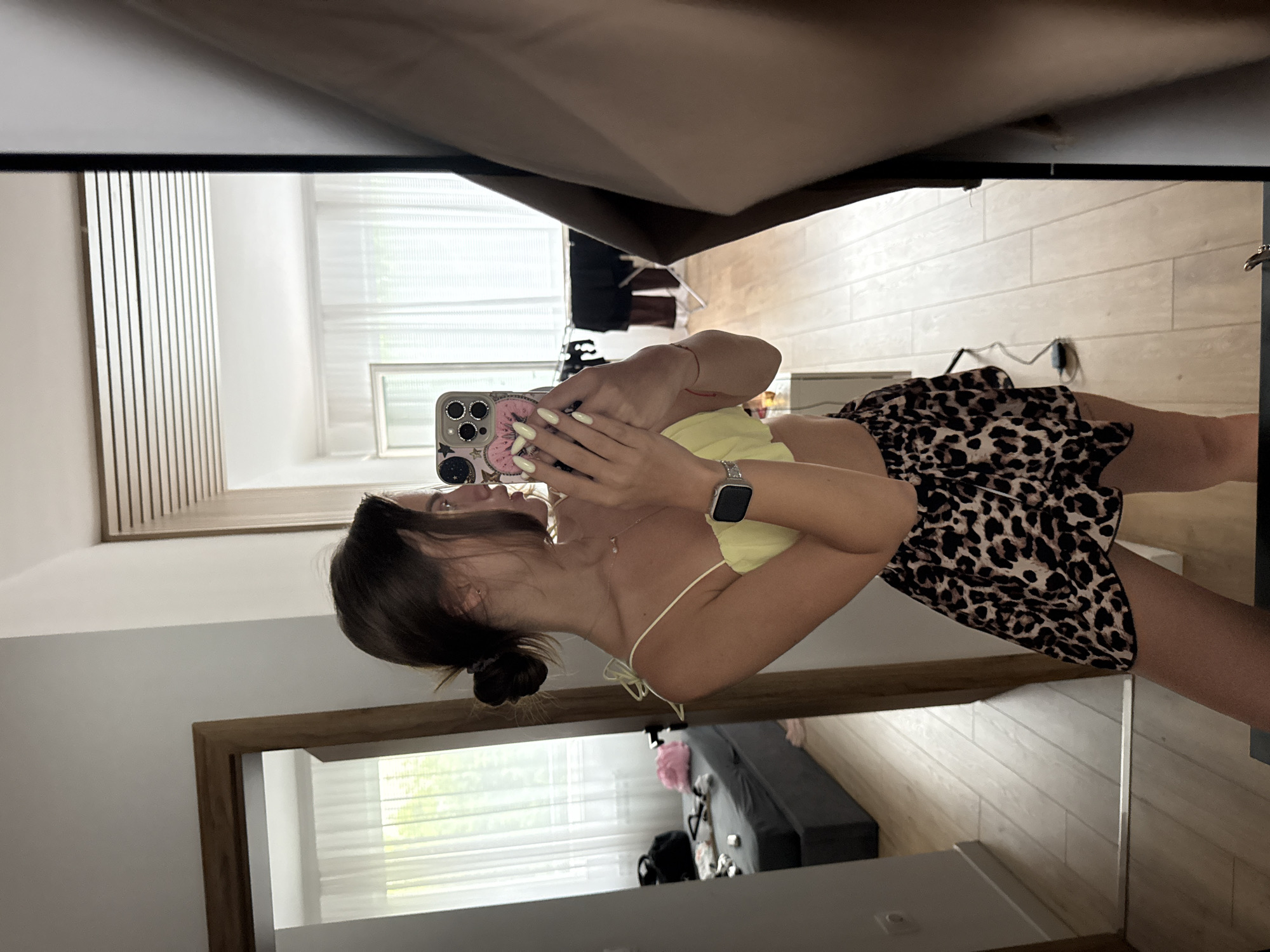 A young woman with her hair in a bun is taking a mirror selfie in a room with light wood flooring. She is wearing a yellow crop top and a leopard-print skirt. In the reflection, a bed with dark grey bedding and a pink pillow is visible in an adjacent room, along with sheer white curtains and a window. The overall setting appears to be a residential apartment or hotel room in Plovdiv, Bulgaria.