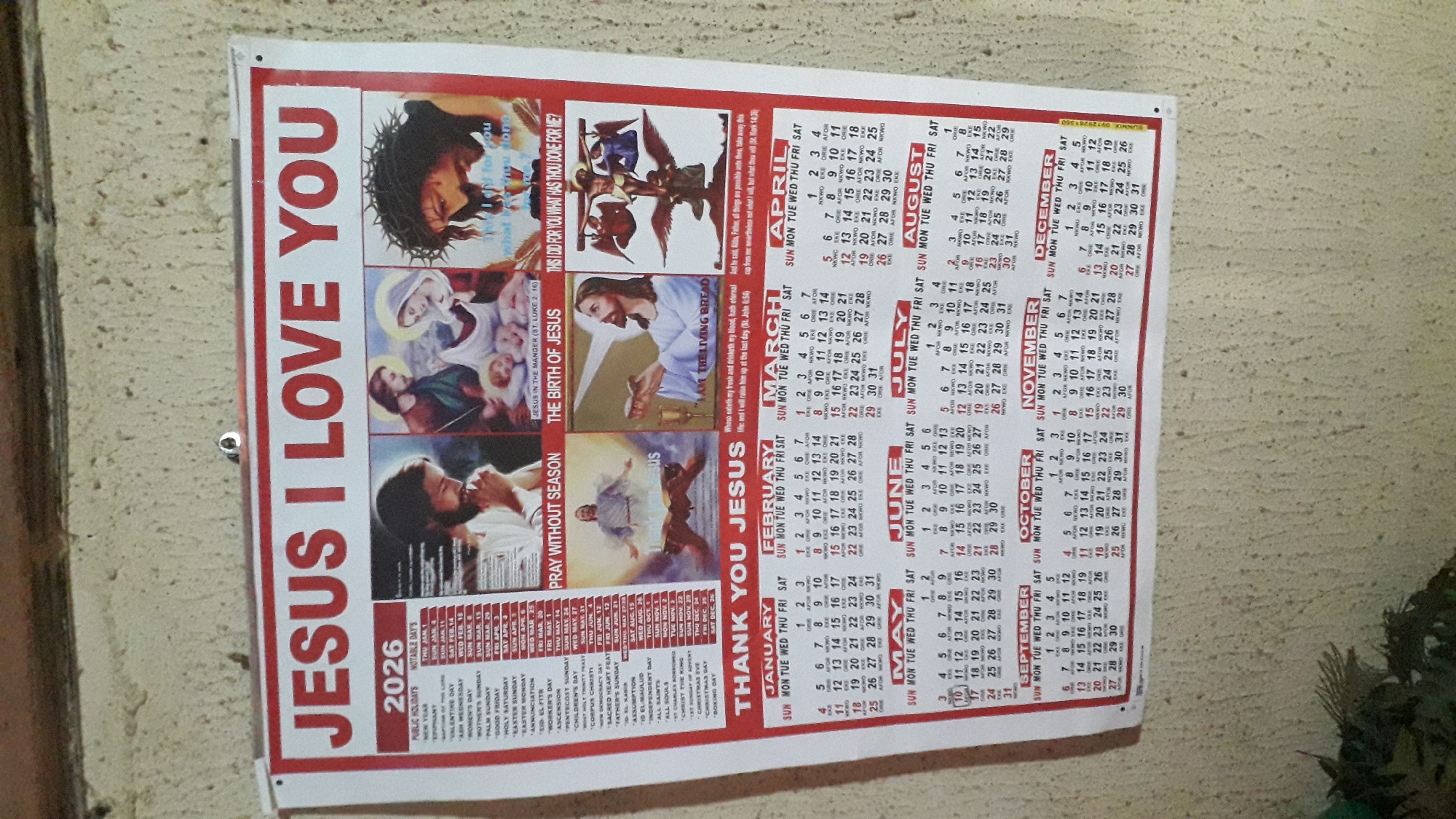 A calendar hangs on a wall, likely inside a building. The calendar prominently features images of Jesus and religious themes. The text "JESUS I LOVE YOU" is printed in large, red letters. Smaller text lists public holidays and notable days for the year 2026. Calendar grids for each month of the year are visible. Small text in the calendar grid indicates that the calendar is for Nkpor, Nigeria.