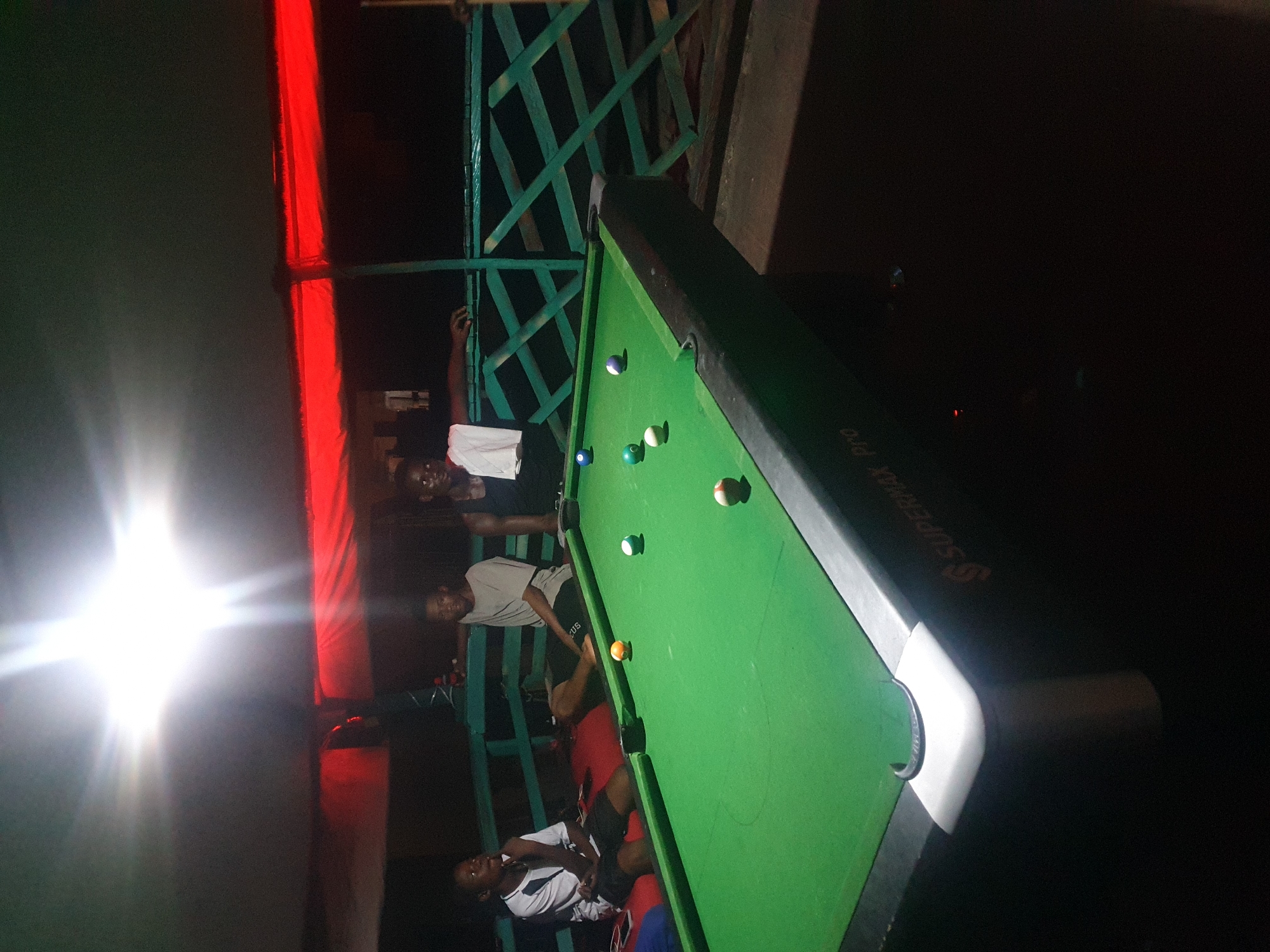 In Lokoja, Nigeria, three young men are gathered around a pool table. The setting appears to be an outdoor recreational area, possibly a bar or lounge, characterized by a green metal railing and a red tarp overhead, illuminated by bright lights. The pool table itself is green felt with various colored balls scattered across it, including a cue ball. One man, wearing a white and black shirt, is seated to the left, engaged in conversation. Another man, in a grey t-shirt, sits to the right of the table, looking towards the camera. The third man, in a black tank top, stands behind the table, leaning on the railing. The lighting is dramatic, with a strong overhead spotlight casting shadows. The overall atmosphere suggests a casual, social gathering.